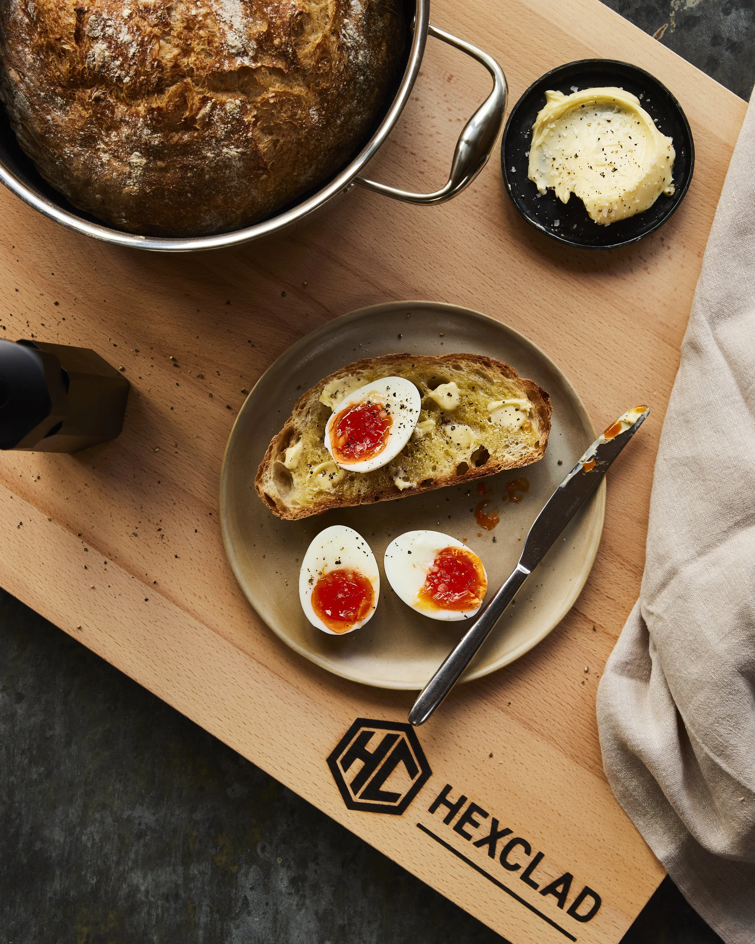 A loaf of crusty bread on a wooden board, with half a slice on a beige plate topped with soft-boiled eggs and sauce. Two halved soft-boiled eggs are on the plate. A small dish of butter and a bread knife are also on the board.