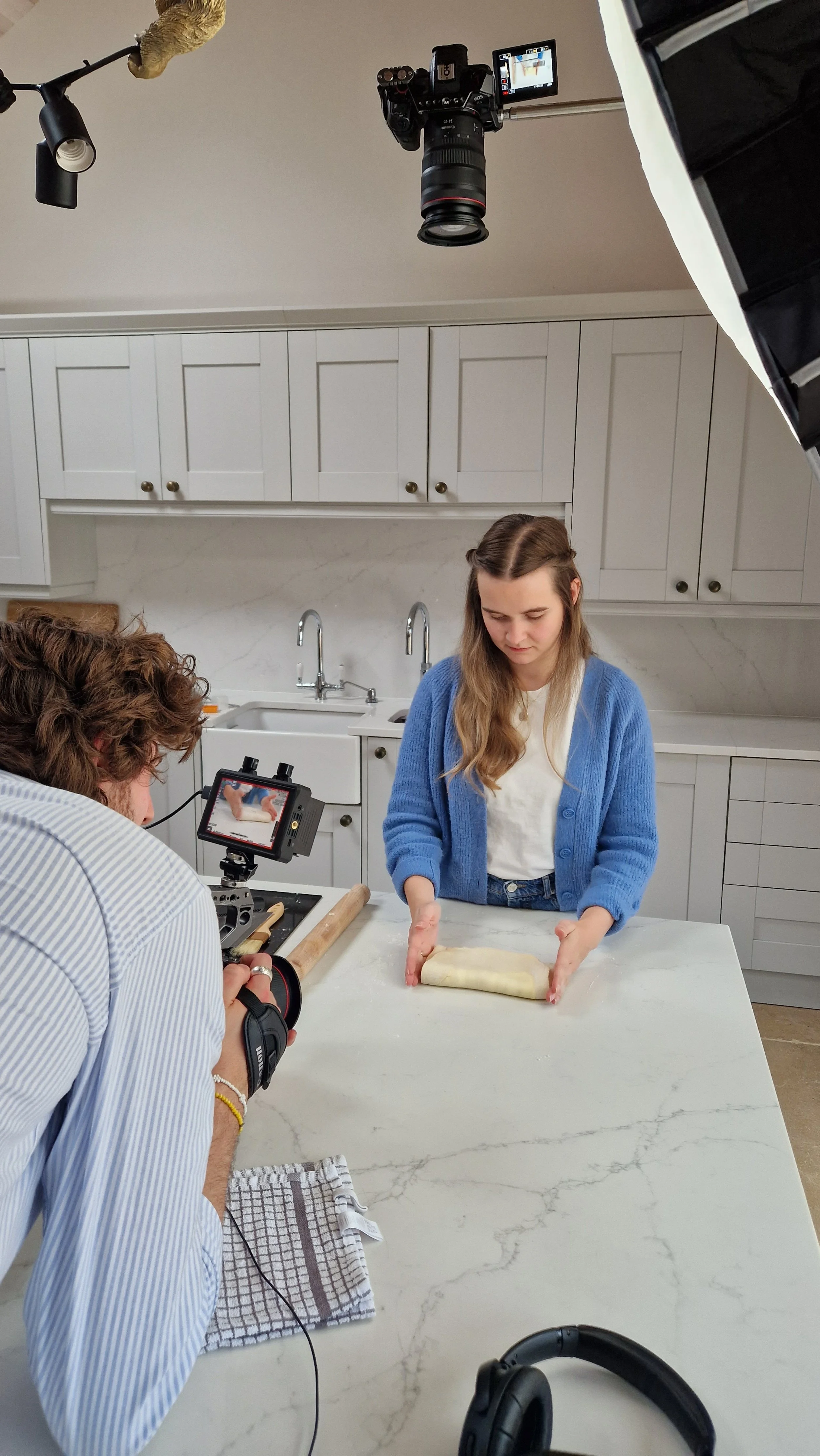 A young woman is filming a cooking scene in a kitchen. She is rolling out dough on a white marble countertop while a camera operator captures the shot. Studio lights and cameras are set up in the space.