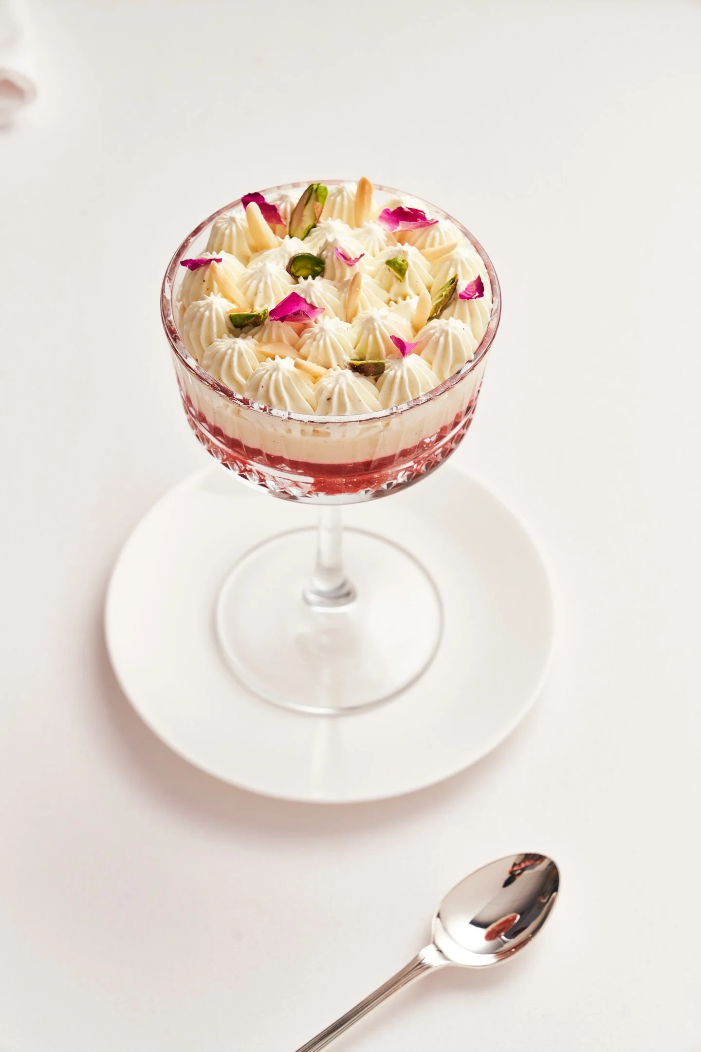 A dessert in a glass bowl decorated with piped cream, pink flower petals, and green pistachio pieces, placed on a white saucer with a silver spoon beside it on a white surface.