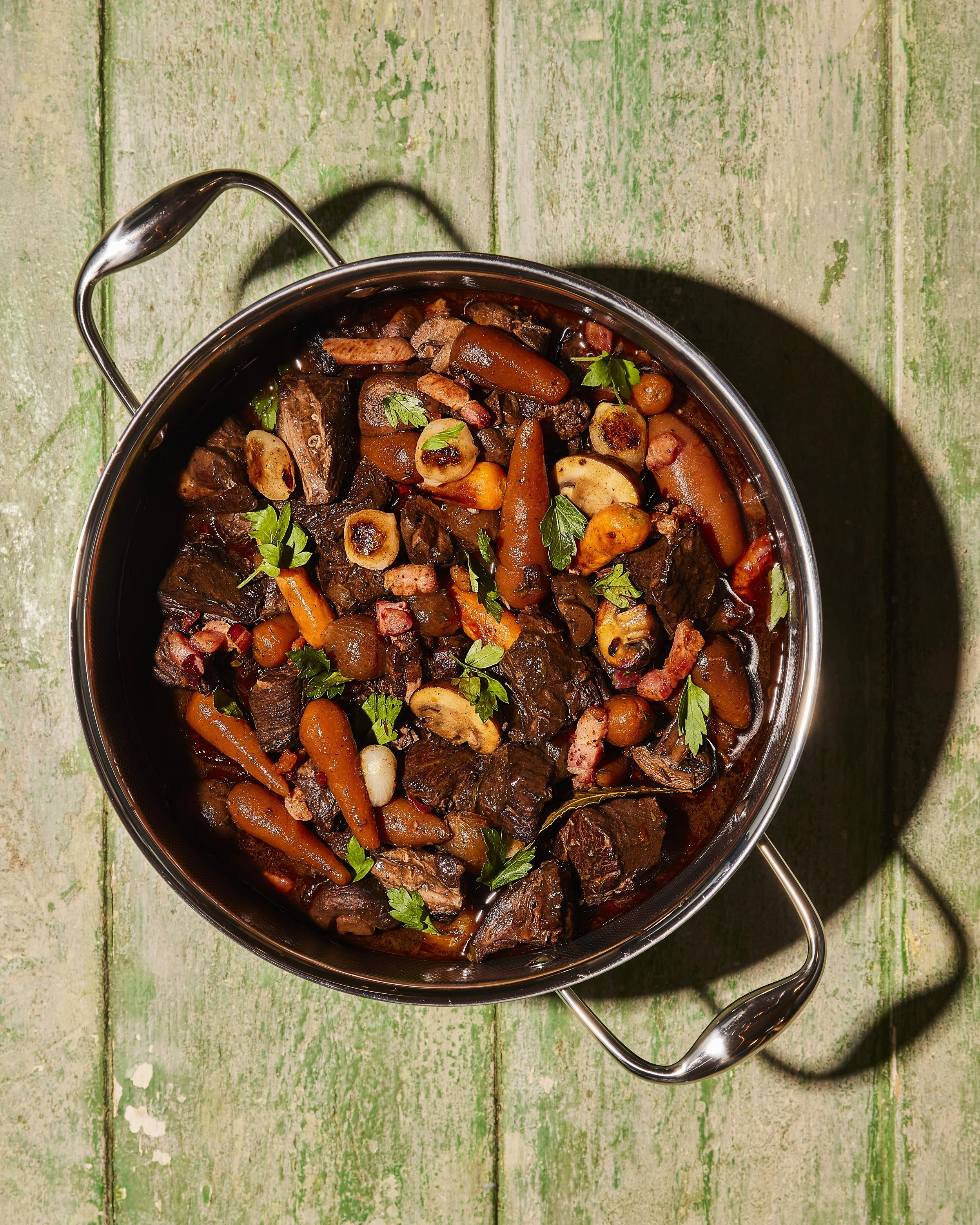 A pot of cooked beef stew with carrots, mushrooms, and parsley on a green wooden surface.