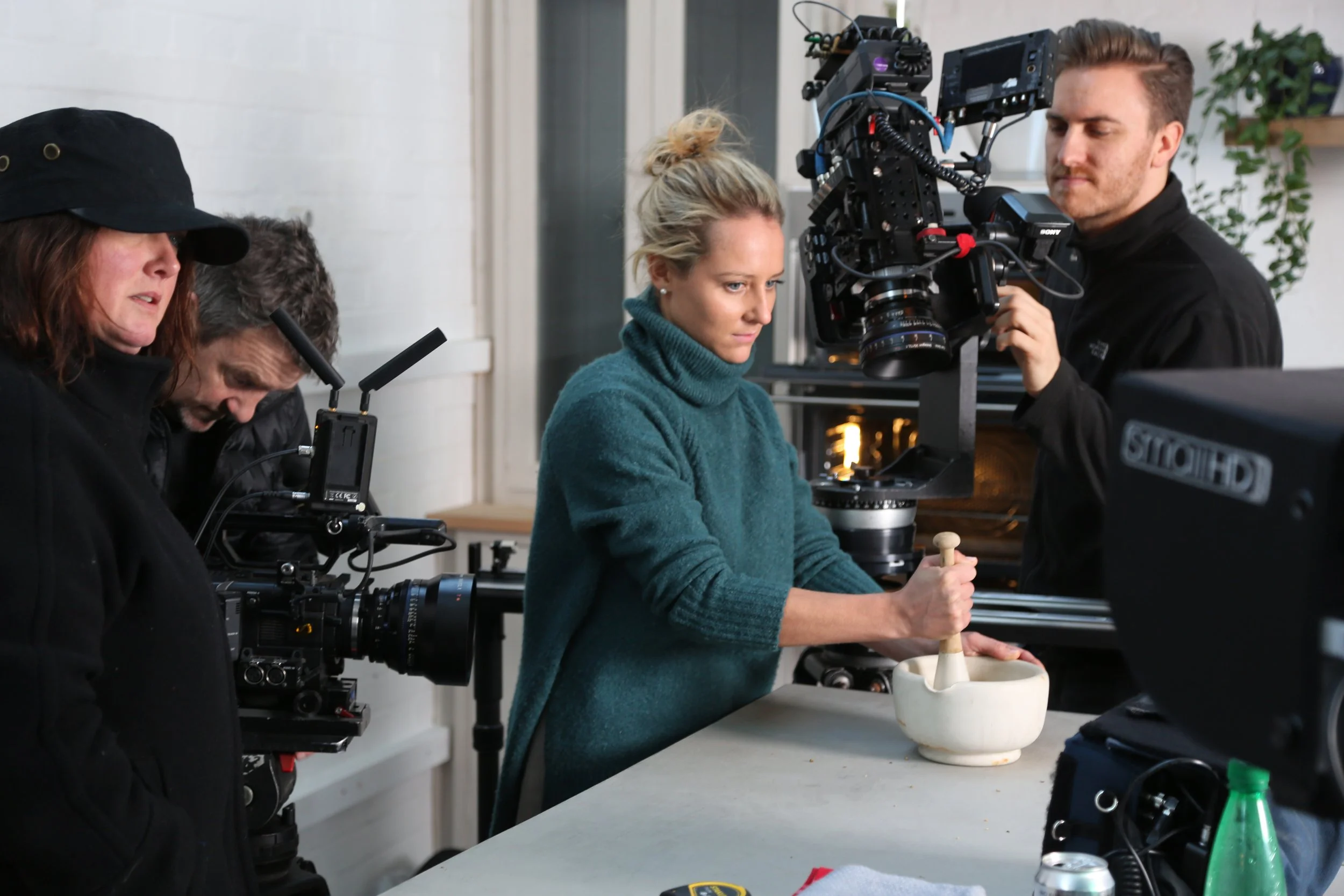 A film crew on set filming a woman stirring a mortar and pestle at a table.