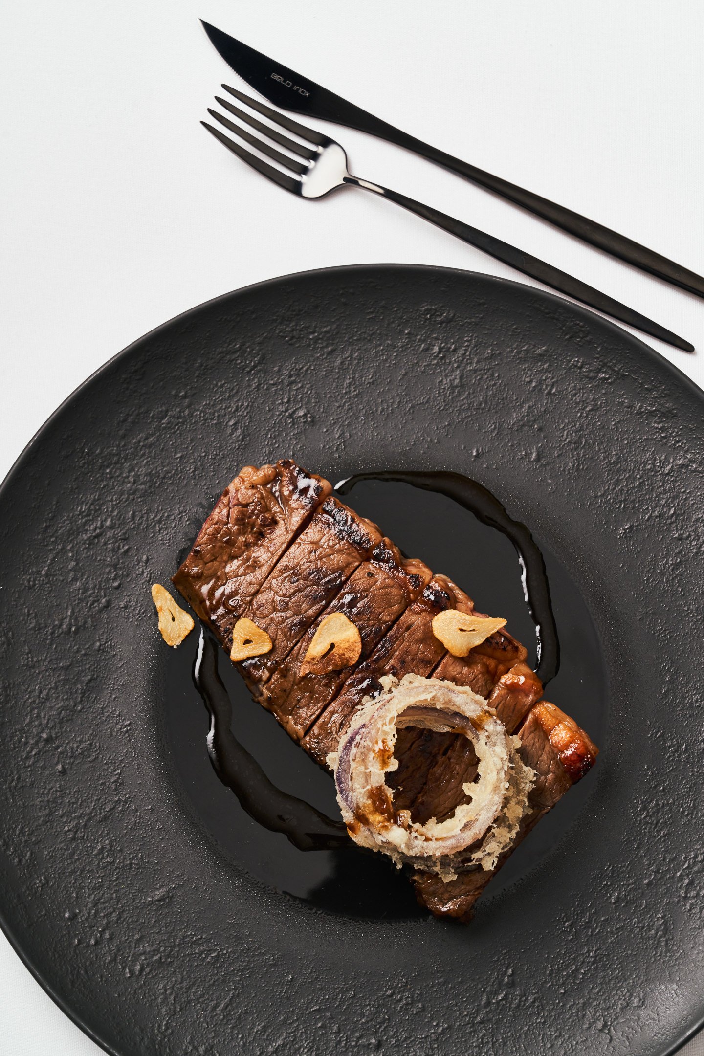 A plated cooked beef short rib with garlic slices and sauce on a black plate, with a black-handled fork and knife on a white background.