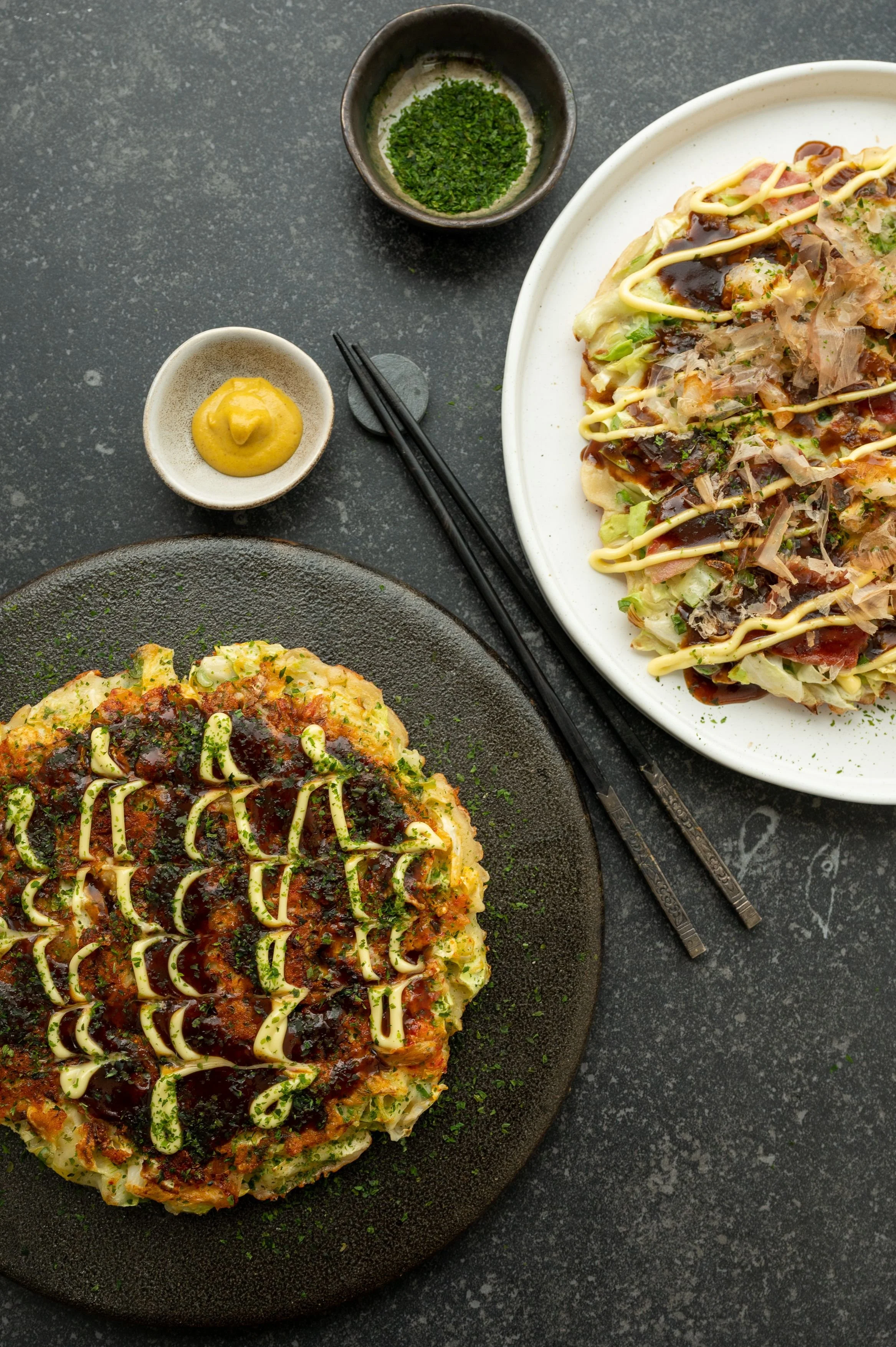 Two okonomiyaki pancakes topped with mayonnaise, teriyaki sauce, and chopped herbs, served on black and white plates with side condiments and chopsticks.