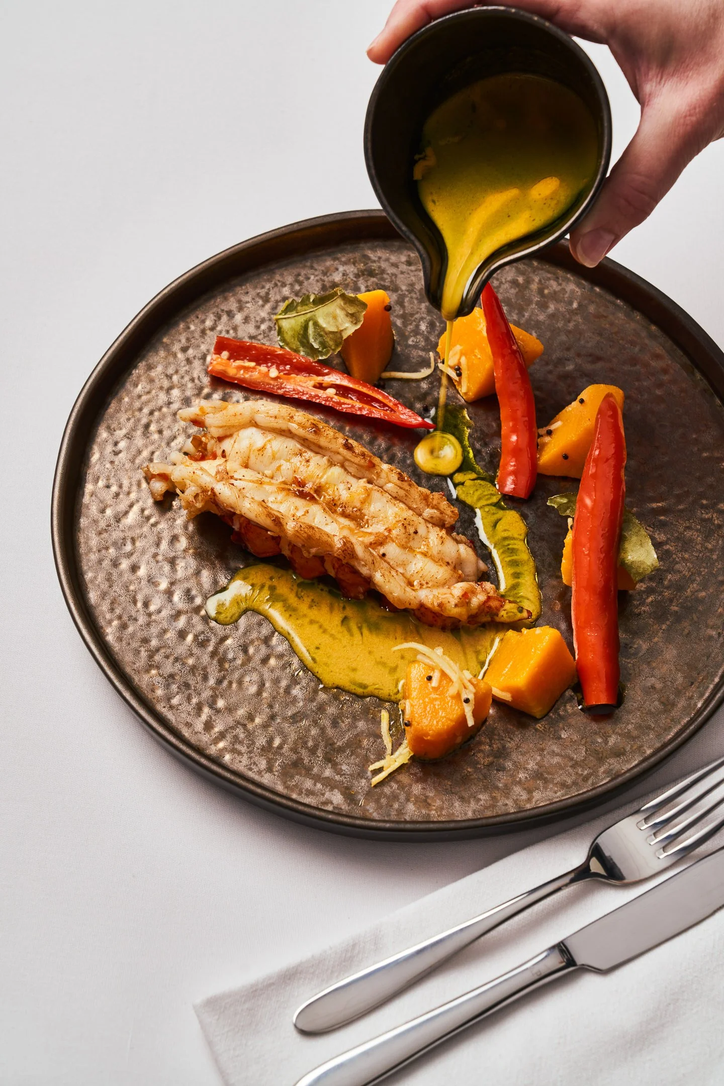 A plate with grilled lobster tail, colorful roasted vegetables, and drizzled green sauce, with a hand pouring yellow dressing.