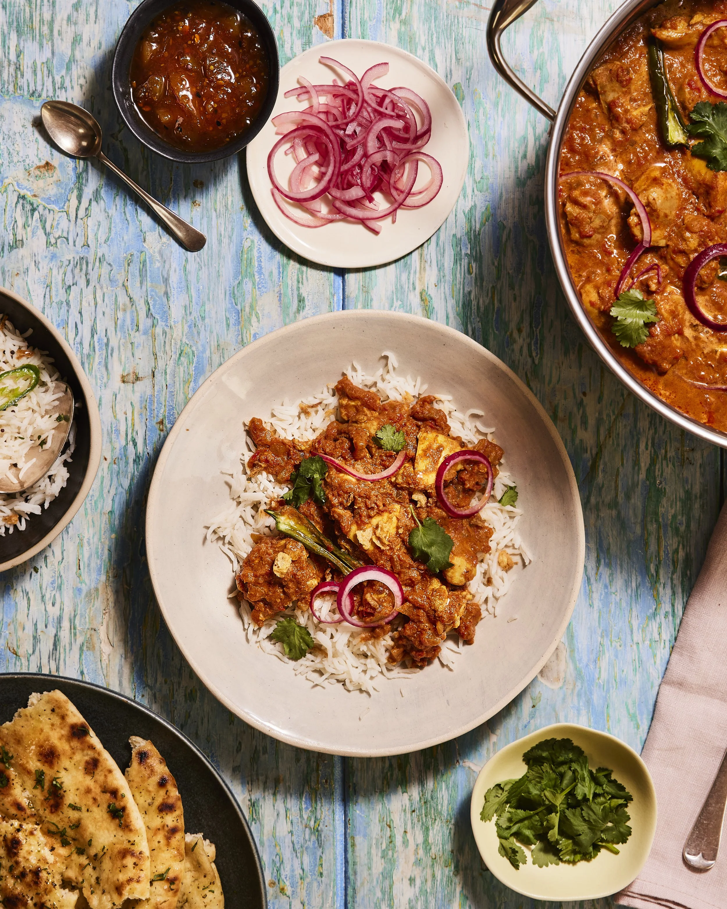 Indian meal with a bowl of curry, plate of rice with curry, naan bread, sliced onions, cilantro, and a small bowl of chopped cilantro on a rustic blue wooden table.