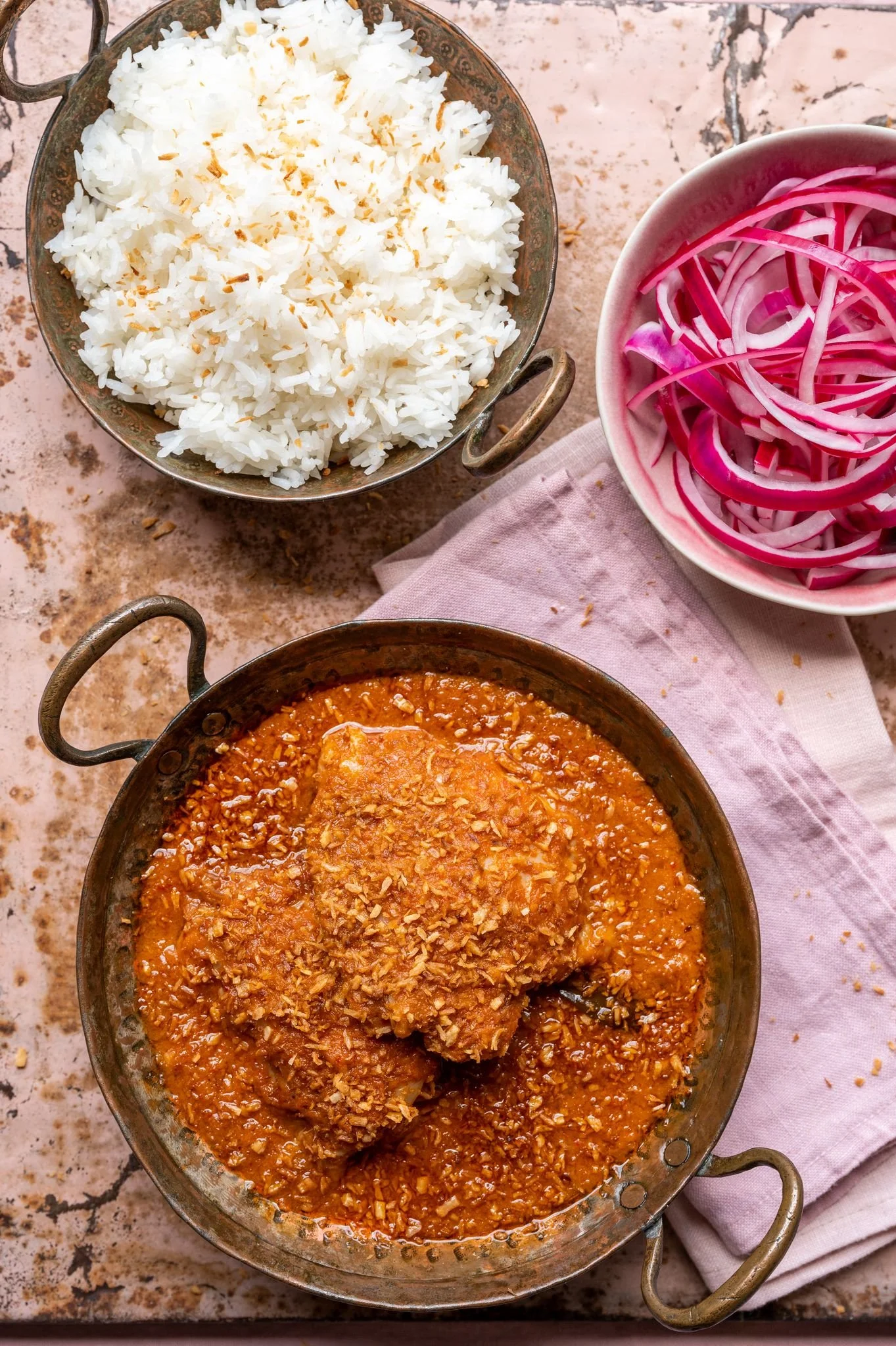 A rustic table setting with a bowl of cooked white rice, a bowl of sliced red onions, and a pan of chicken cooked in a spicy sauce.