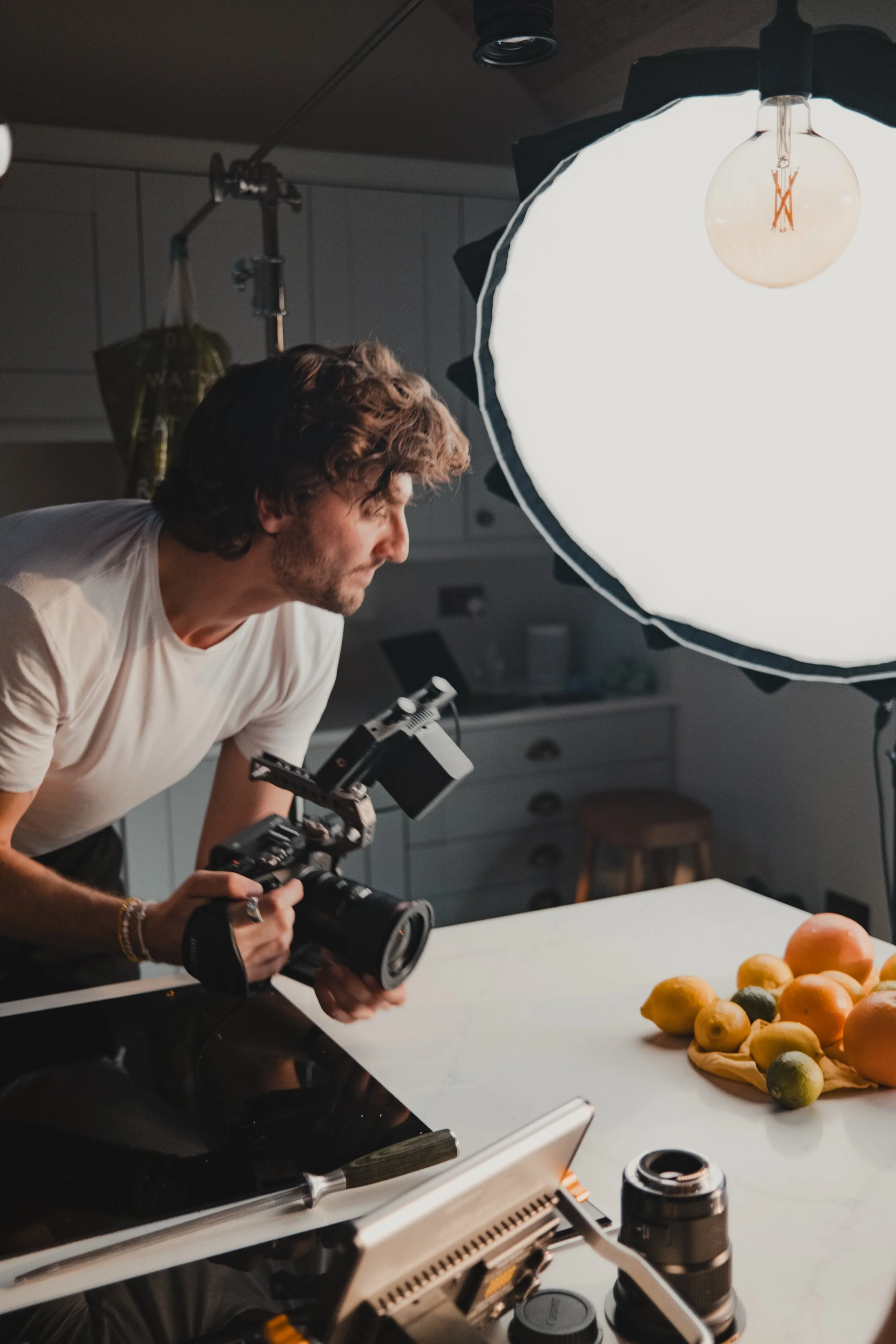 A man is filming a food photography shoot in a studio, with a large white diffuser light overhead. On the table, there are various fruits, including lemons and grapefruits, arranged on a cloth.