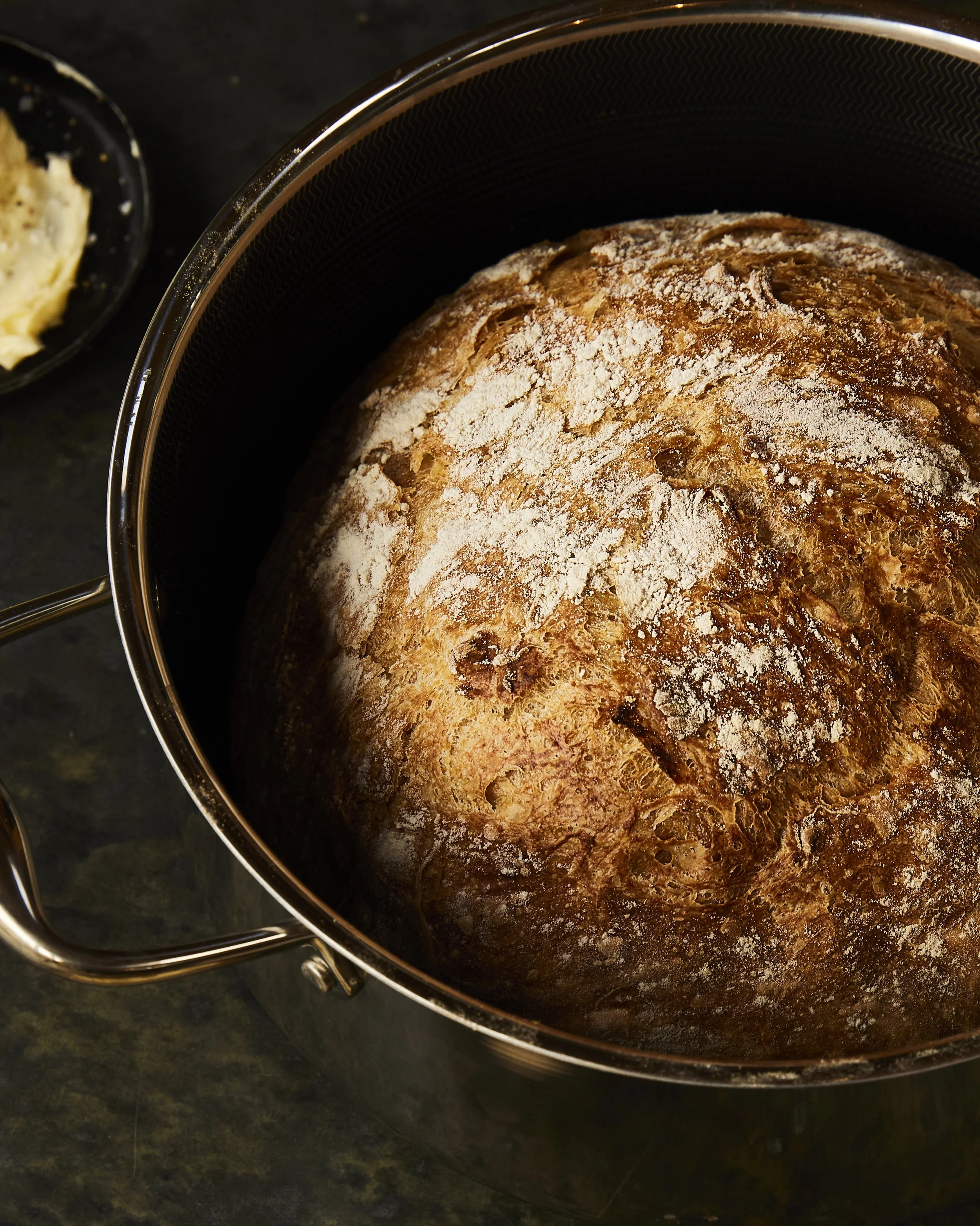 Loaf of bread rising in a baking pot with flour dusted on top, placed on a dark countertop.