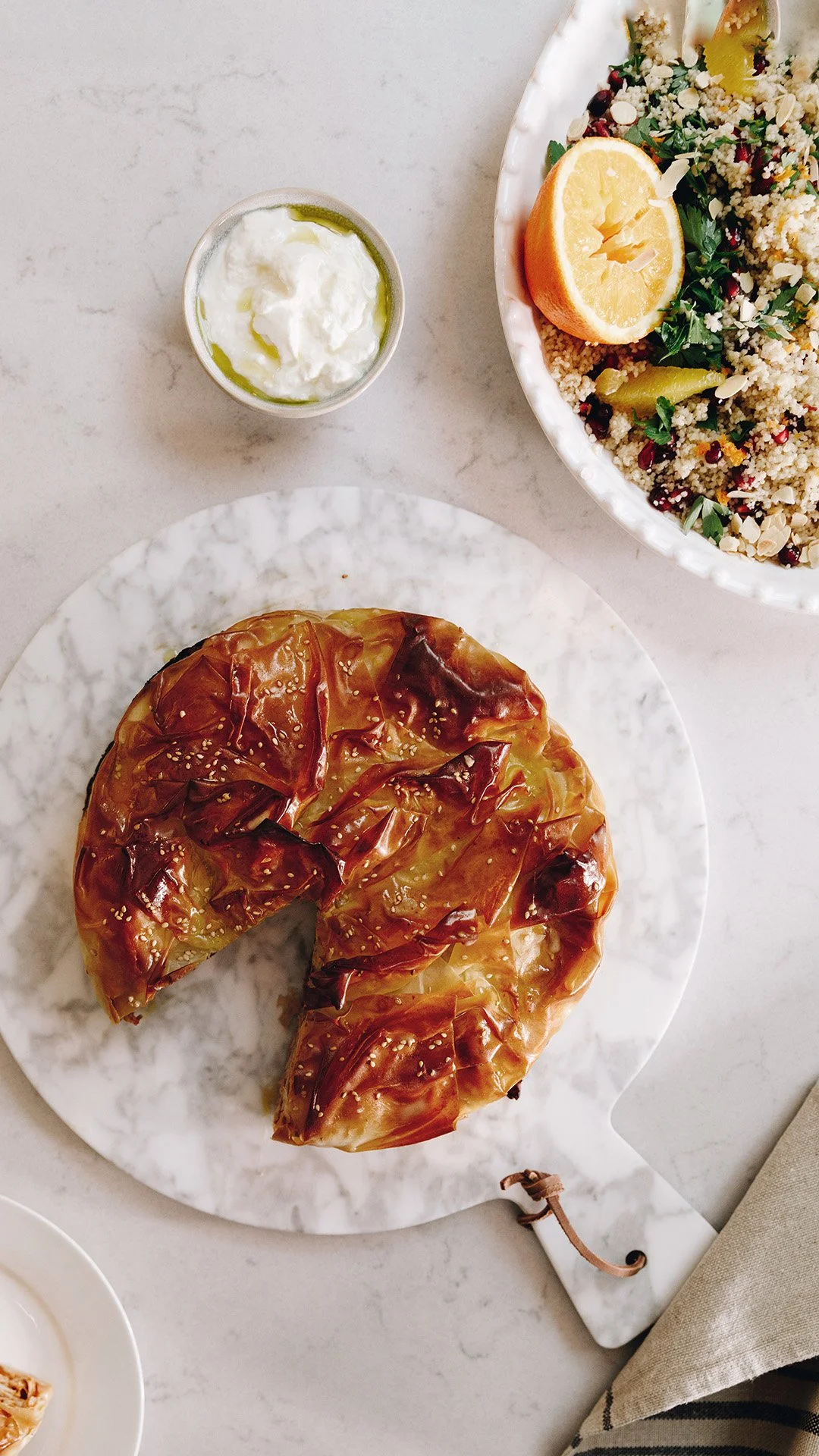 A round pie with a glossy caramelized top on a white marble platter, sliced one piece out, with a small cup of whipped cream or yogurt, a bowl of quinoa salad with lemon wedge, herbs, and vegetables visible, on a white marble table.