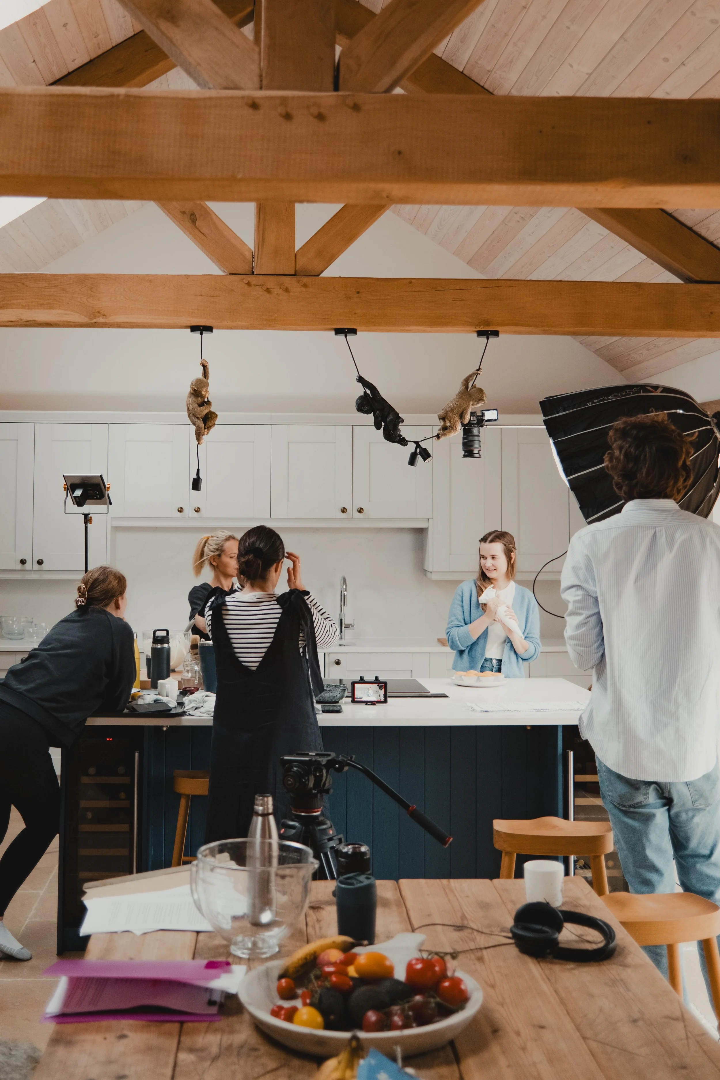 A group of women making a video in a bright, modern kitchen with white cabinets, with a camera crew filming them. Monkeys hanging from the wooden beams overhead.