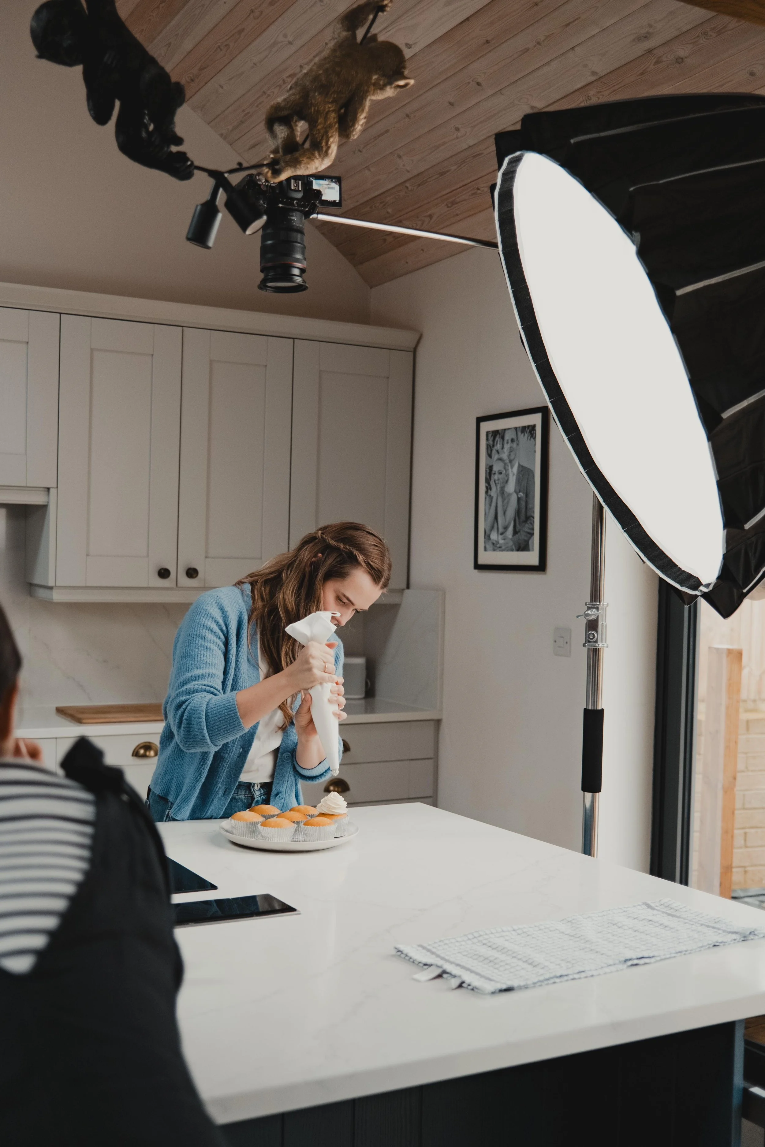 A woman is decorating cupcakes in a kitchen with studio lighting and a black and white framed photograph on the wall.