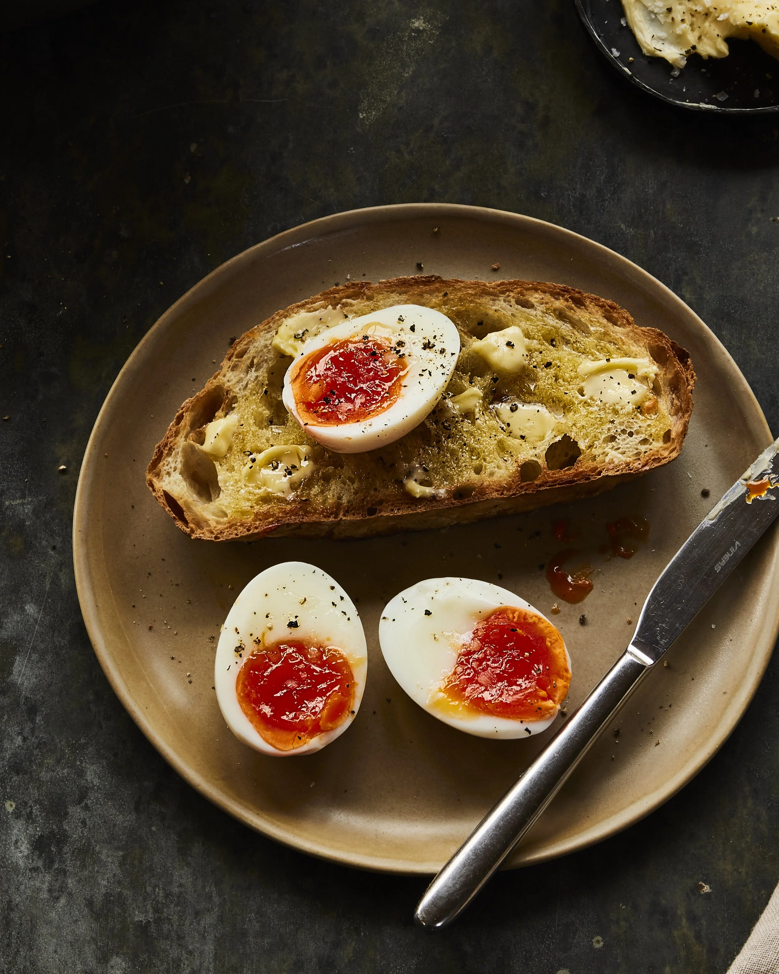 Sliced toasted bread with melted cheese, topped with halved soft-boiled eggs on a beige plate, with a butter knife, on a dark textured surface.