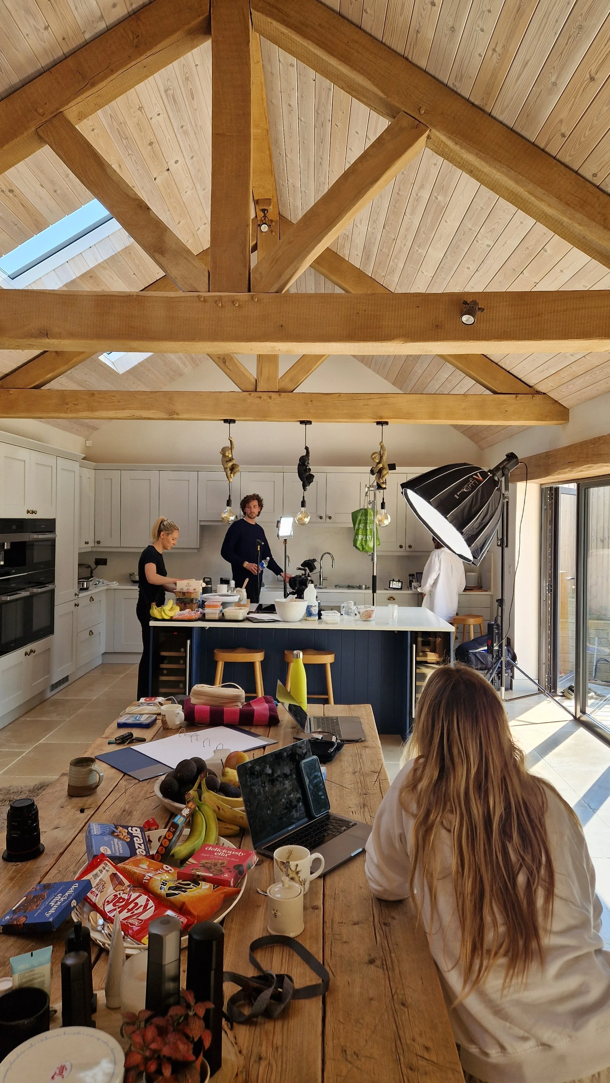 People filming a cooking or food segment in a bright, cozy kitchen with wooden beams and a skylight, cluttered table with snacks and a laptop in the foreground.