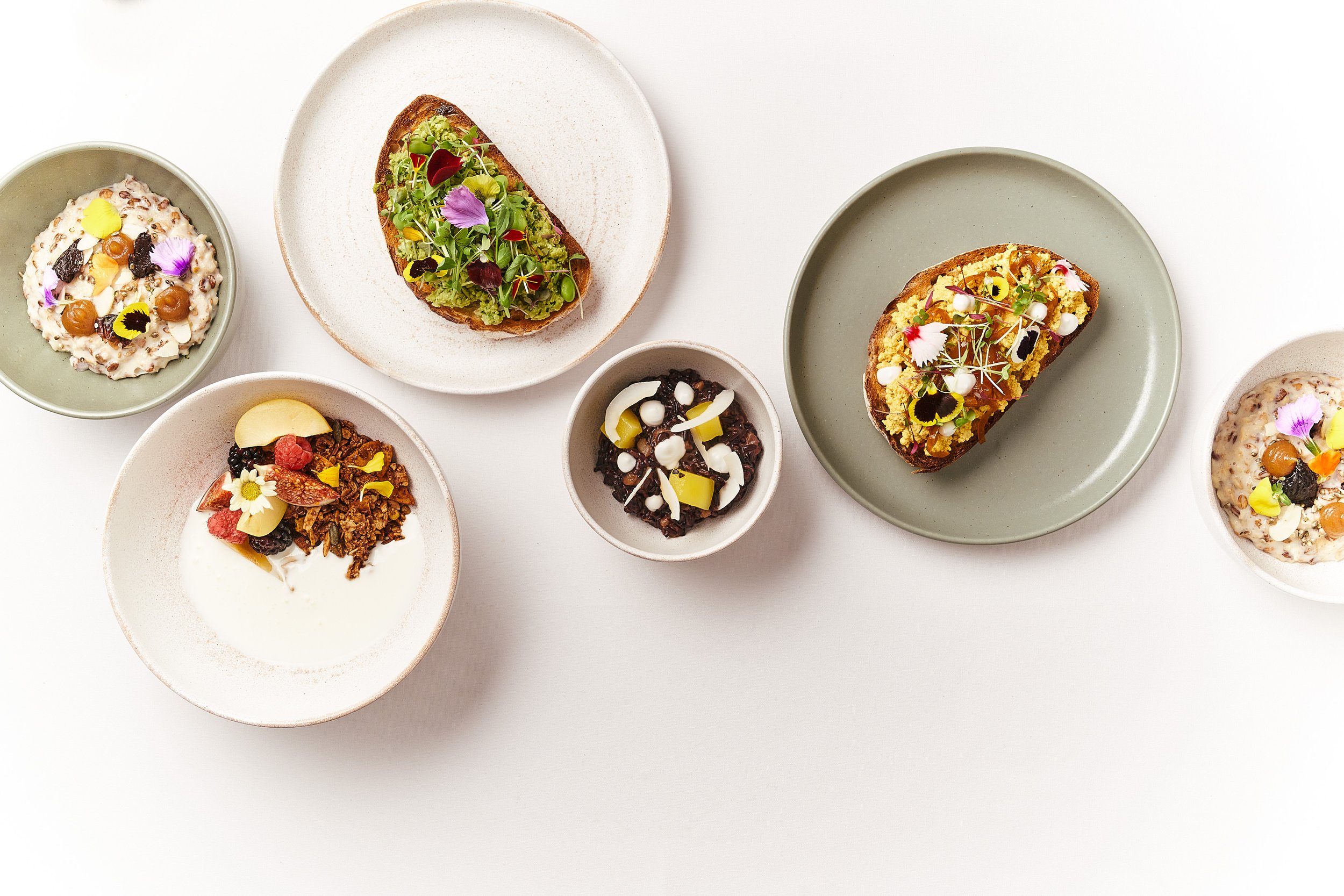 Assorted dishes served on white and gray plates, including small bowls with yogurt, granola, and fruit, along with slices of bread topped with microgreens and edible flowers, displayed on a white surface.