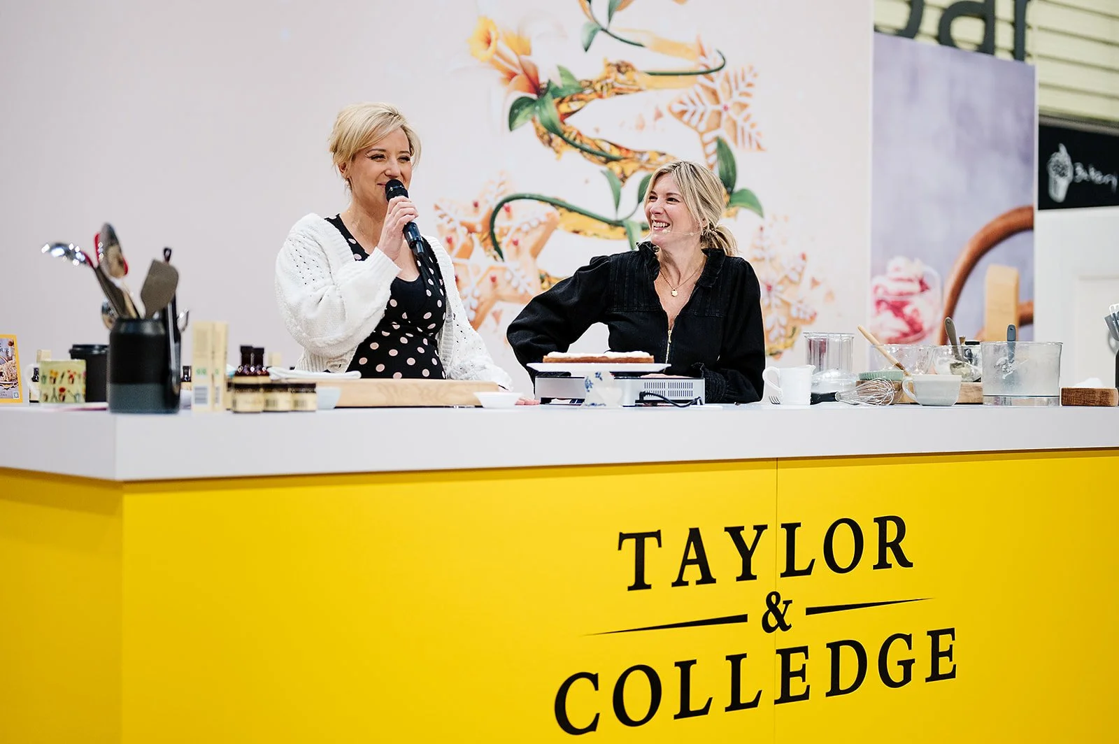 Two women smiling and talking on a yellow booth labeled "Taylor & College," with baking tools and ingredients on the counter in a cooking demonstration or class.