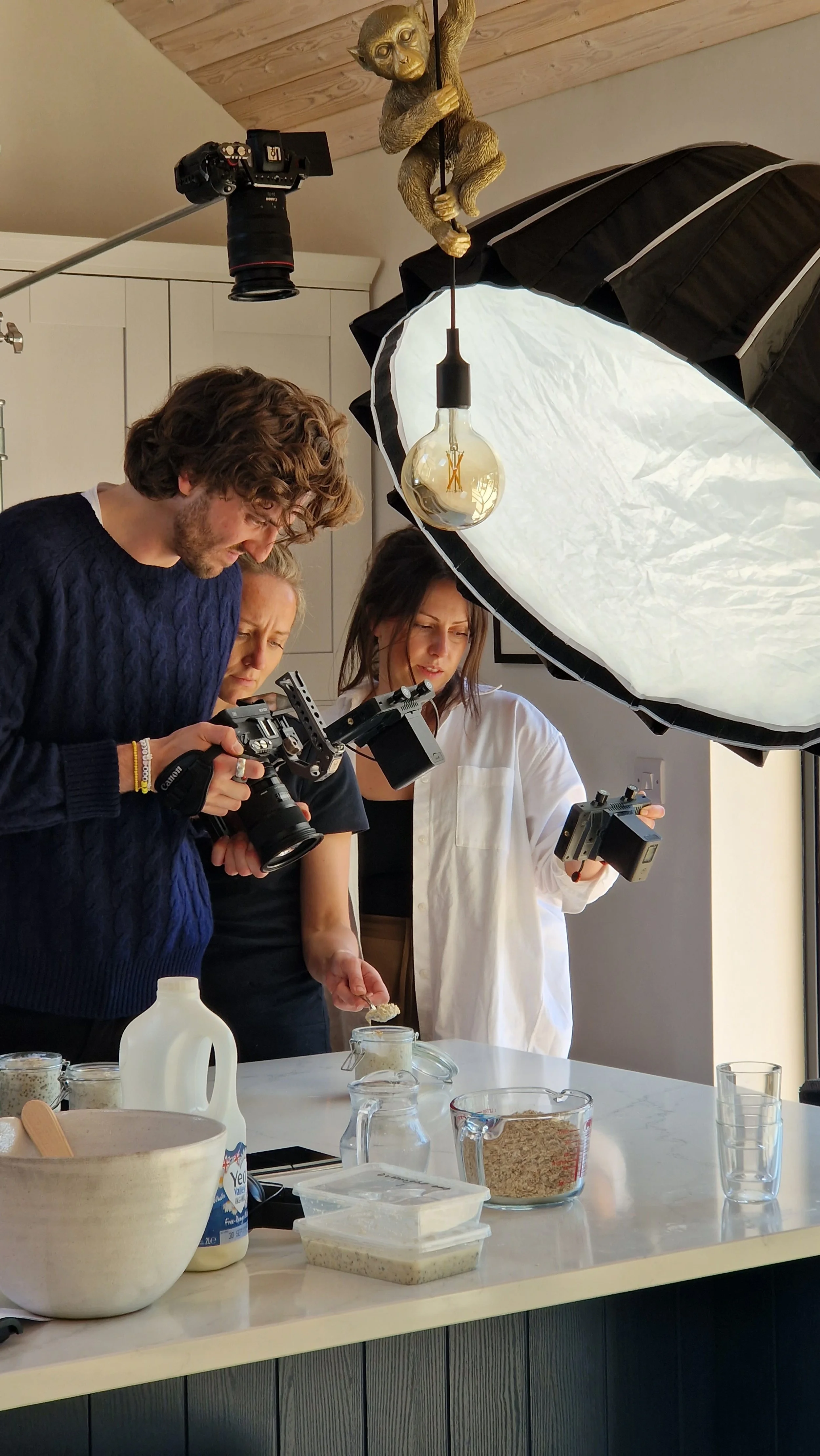 Three people filming a cooking demonstration in a kitchen. One person is holding a camera, another is holding a monitor, and a woman is scooping from containers on a white countertop. The scene is illuminated by professional lighting equipment, inclu