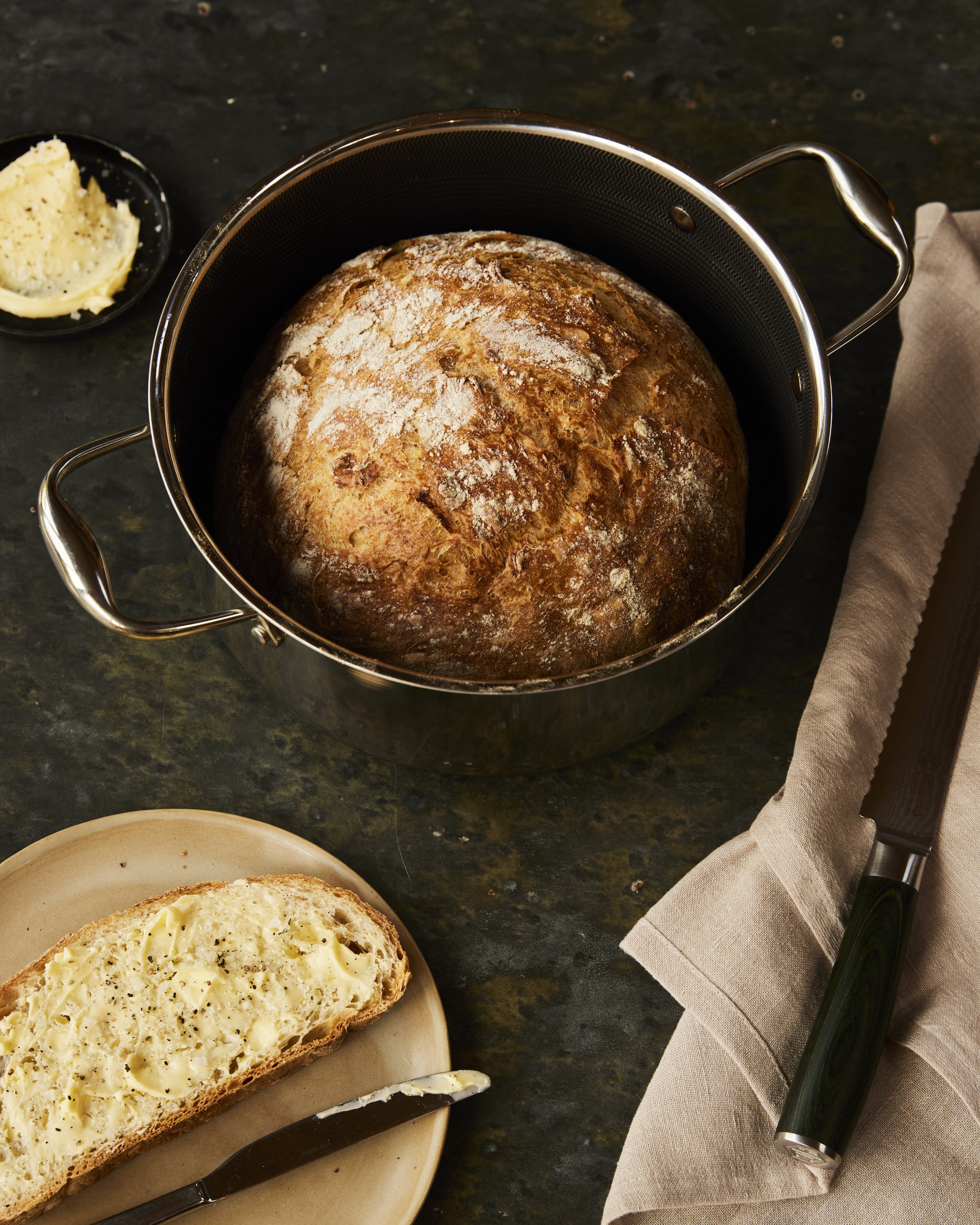 A freshly baked round loaf of bread in a black pot, with a small plate of butter and a slice of bread spread with butter on a beige plate nearby. There is a butter knife on the plate and a bread knife resting on a folded beige cloth napkin to the sid