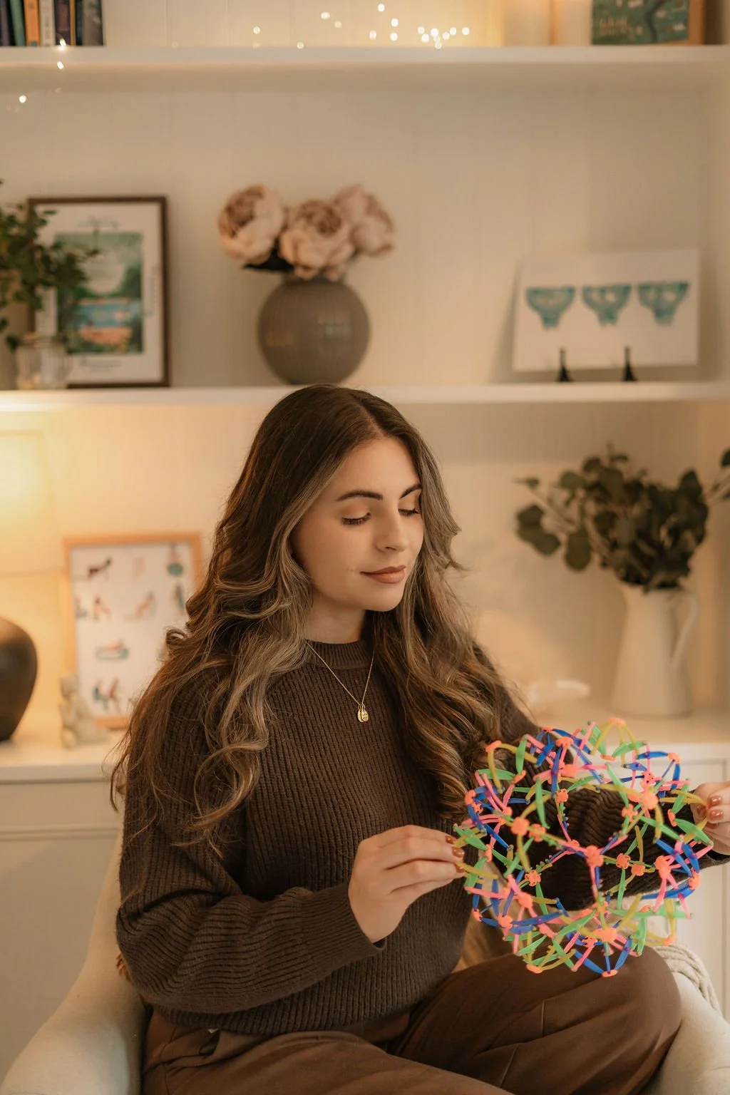 A woman with long wavy brown hair wearing a brown sweater and necklace, sitting on a beige chair, holding colorful slime shaped like a sphere with pinwheel decorations, in a warmly lit room with books, framed pictures, a vase with flowers, and decorative objects in the background.
