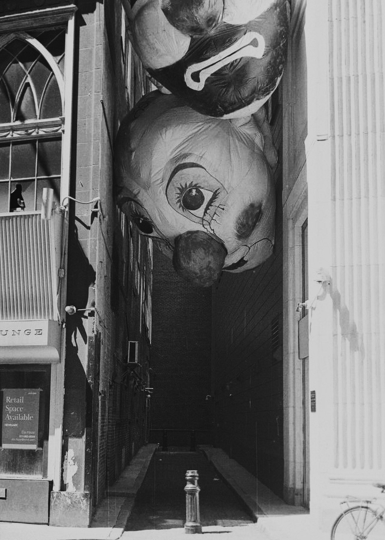 A street scene with a large clown mural painted on the upper part of a building, featuring exaggerated facial features, a big nose, and a painted eye with makeup. The image is in black and white.