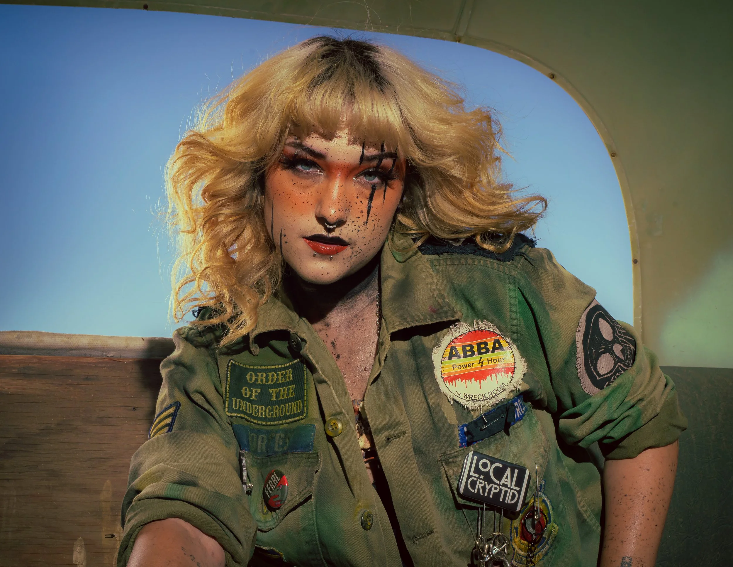 A woman with curly blonde hair and makeup resembling tiger stripes and a painted face wearing a military-style jacket with patches and badges.