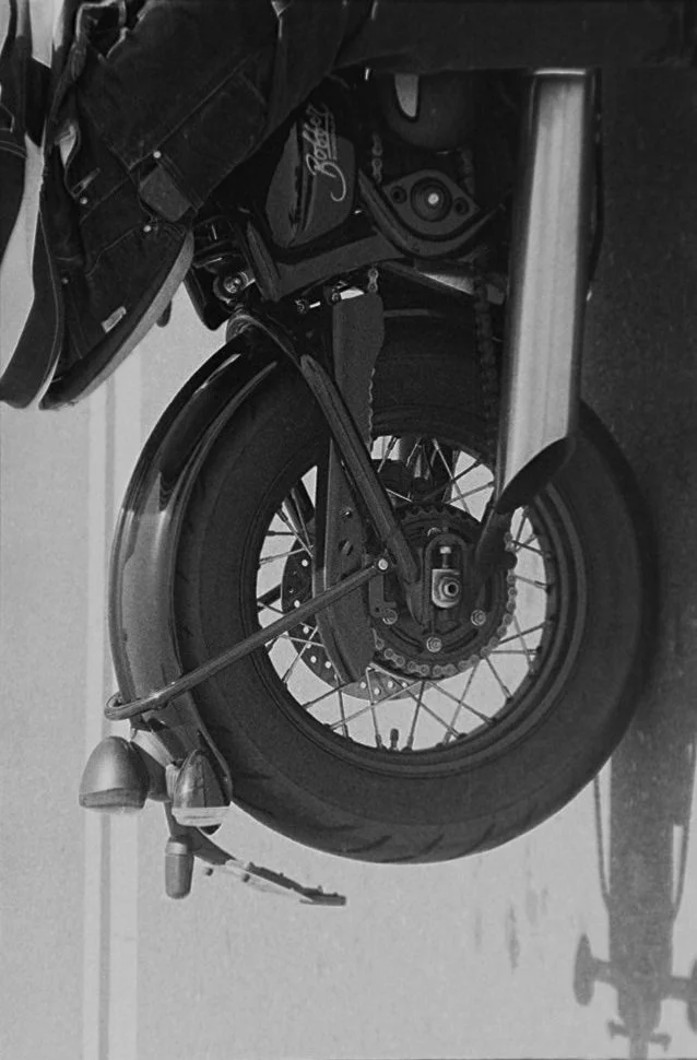 A motorcycle wheel with a tire, disc brake, chain, and part of the frame visible, and a saddlebag attached to the side.