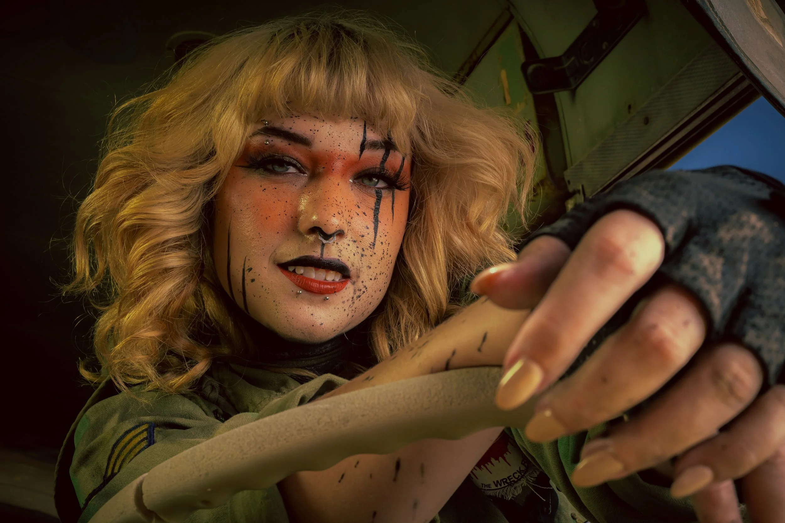 Person with vibrant makeup and messy blonde hair looking directly at camera, holding a steering wheel inside a vehicle.