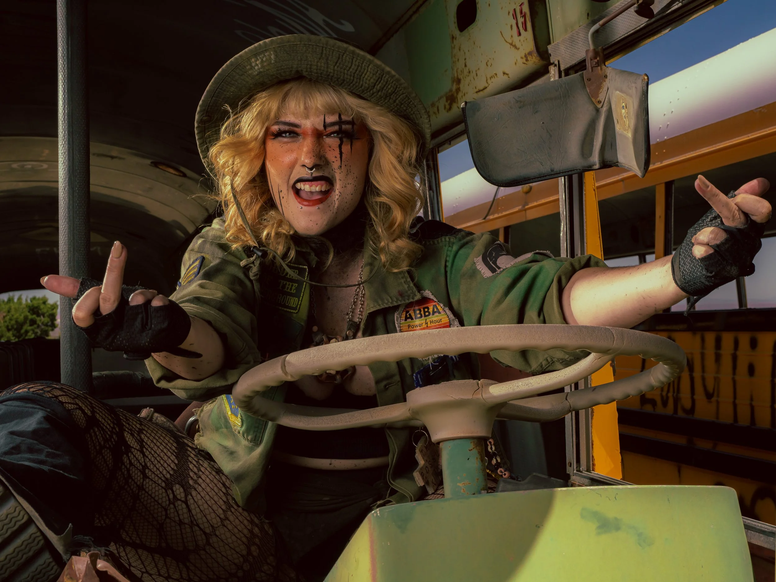 A woman with blonde hair, face paint, and gothic makeup is sitting in the driver’s seat of a school bus. She is wearing a sheriff hat, a green military-style jacket, and fishnet stockings, and making a rebellious hand gesture.