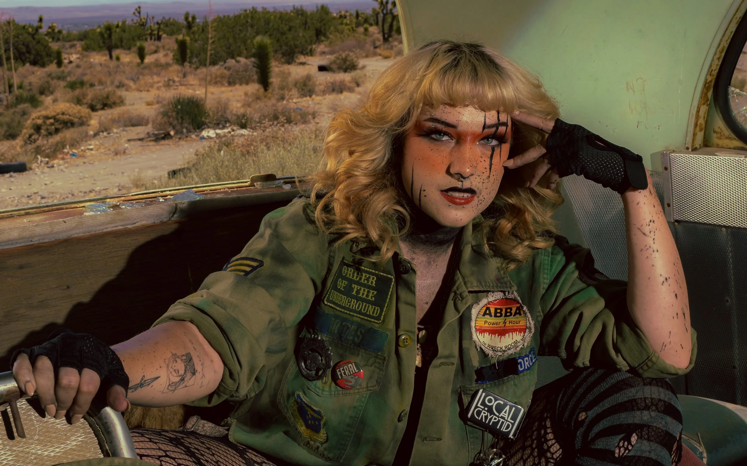 A woman with blonde curly hair, wearing a green military jacket decorated with patches, is sitting in an open vehicle in a desert landscape. She has heavy makeup with black paint streaks on her face and arm, and is wearing a black glove on her right 