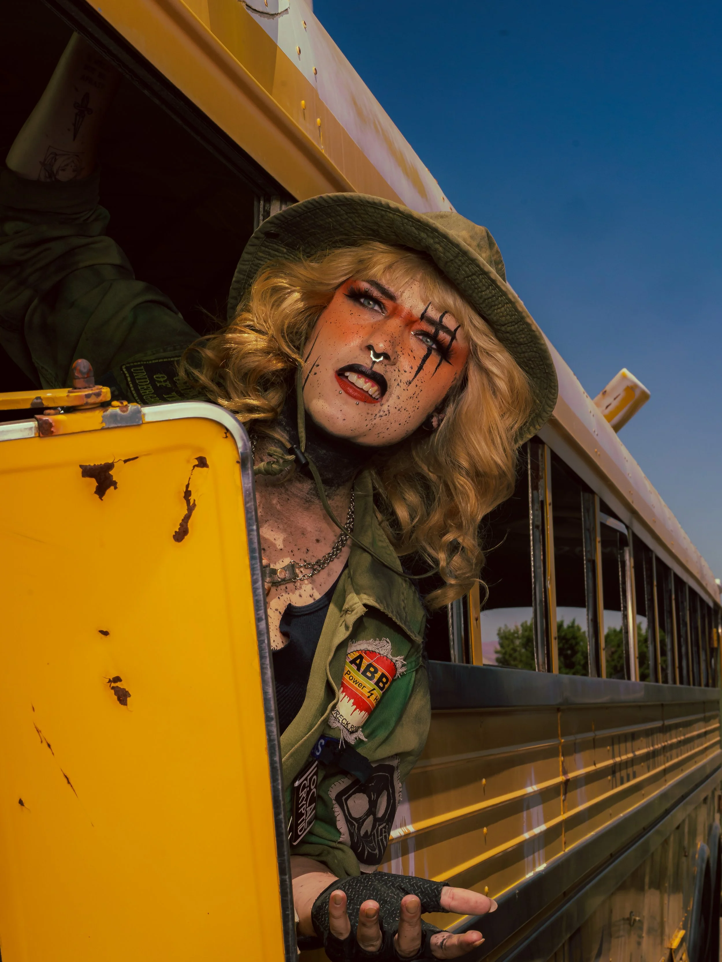 A person with curly blond hair, painted face, and camouflage outfit leaning out of a yellow school bus door, under a clear blue sky.