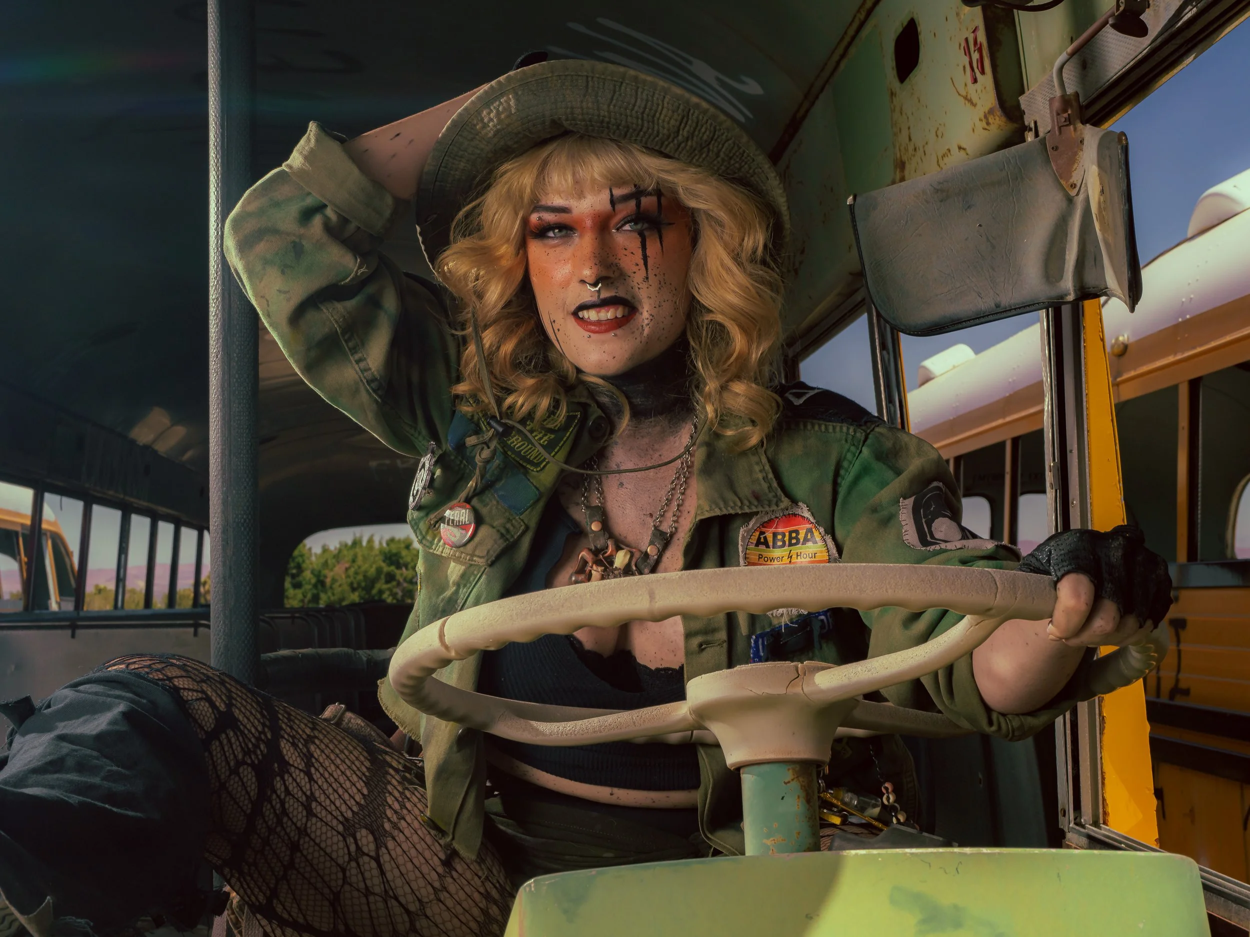 A woman with long blonde curly hair, makeup, a septum piercing, black fishnet stockings, and a camouflage jacket sitting in a vehicle, holding the steering wheel, posing with a confident expression, and wearing a large hat.