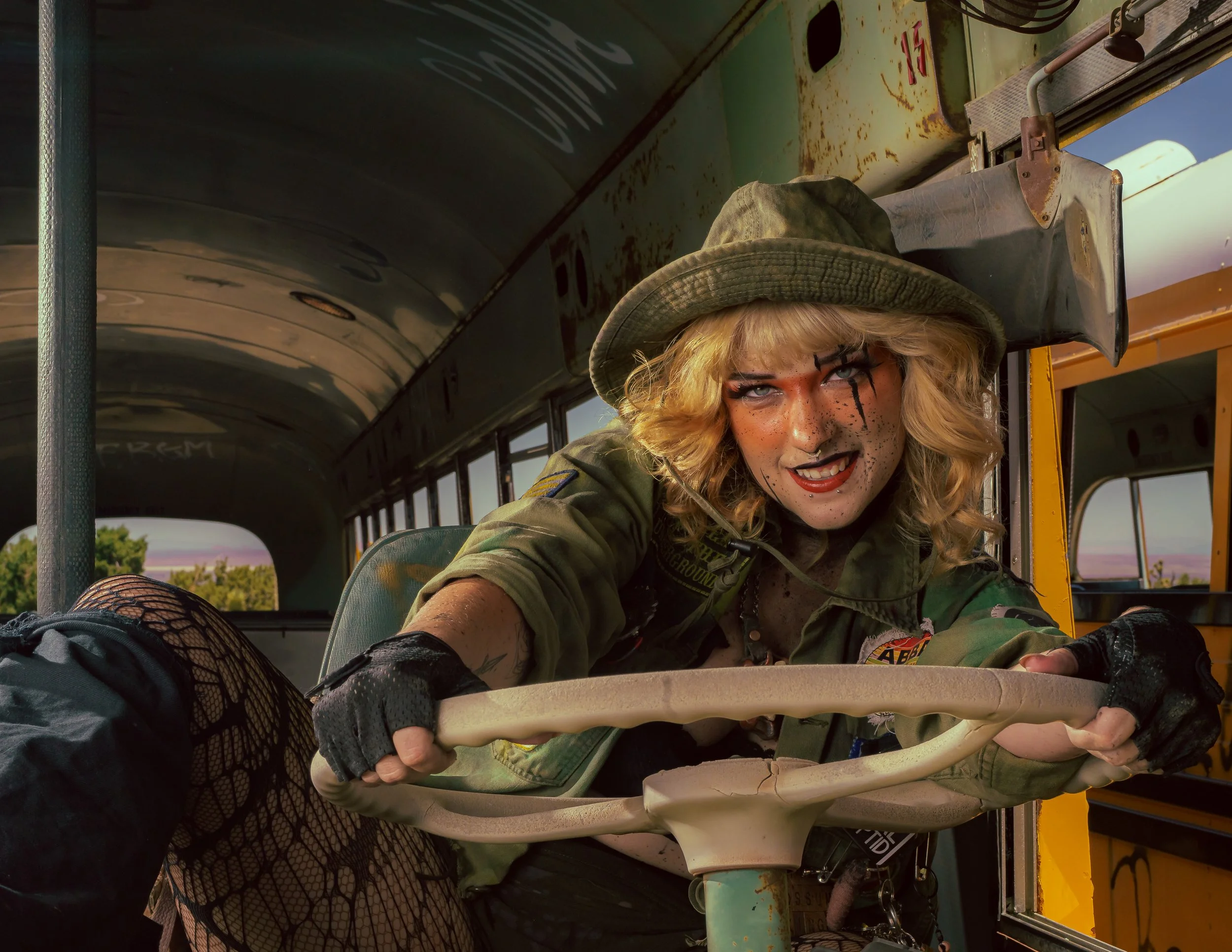 A woman dressed as a creepy, punk schoolgirl leaning out of a yellow bus, with messy blonde hair, dark makeup, and a military jacket, holding onto the bus's steering wheel, smiling mischievously.