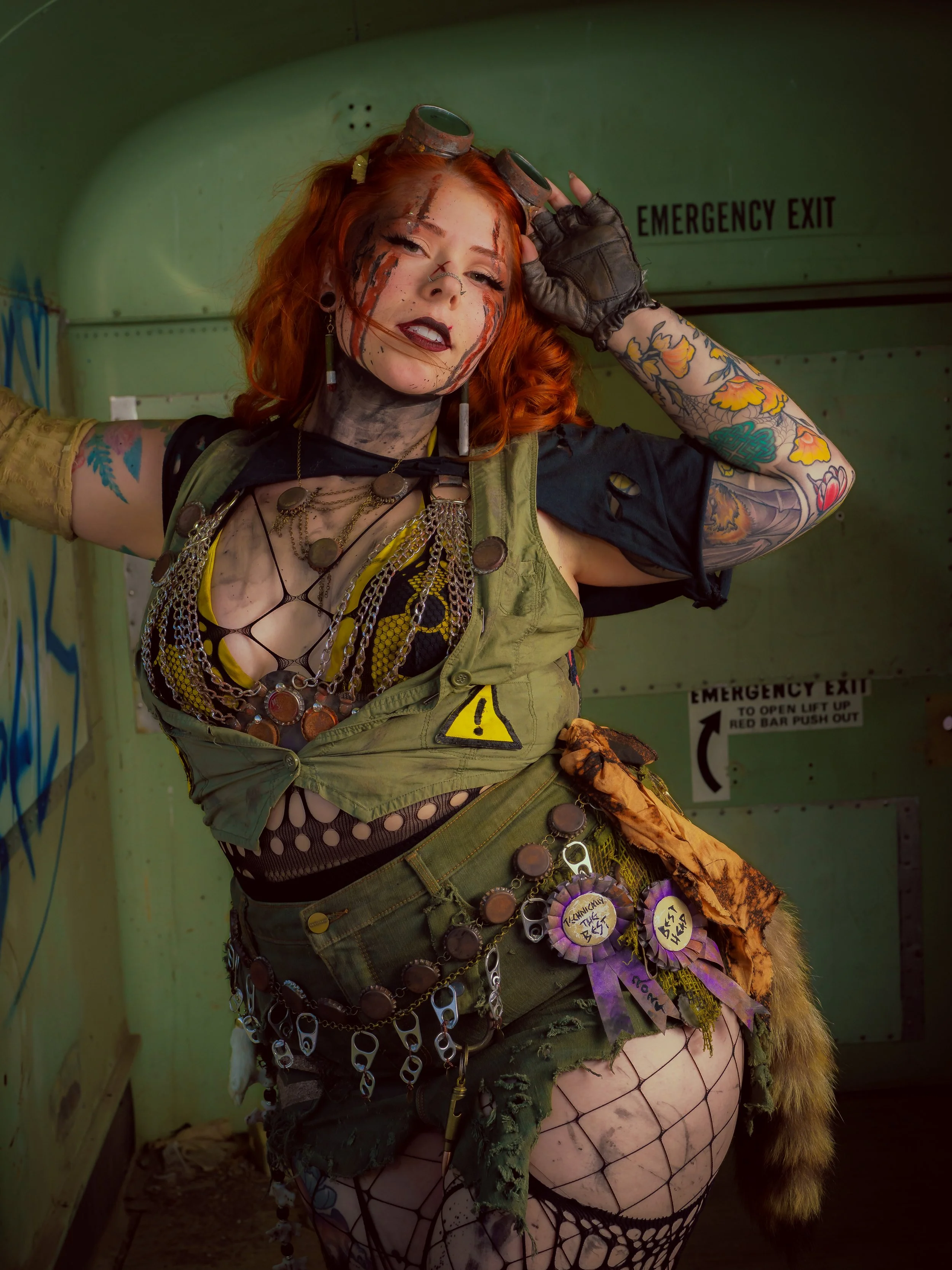 A woman with red hair and makeup in a post-apocalyptic or punk style in front of an emergency exit door, dressed in a ripped, camouflage outfit with chains, pins, and tattoos.