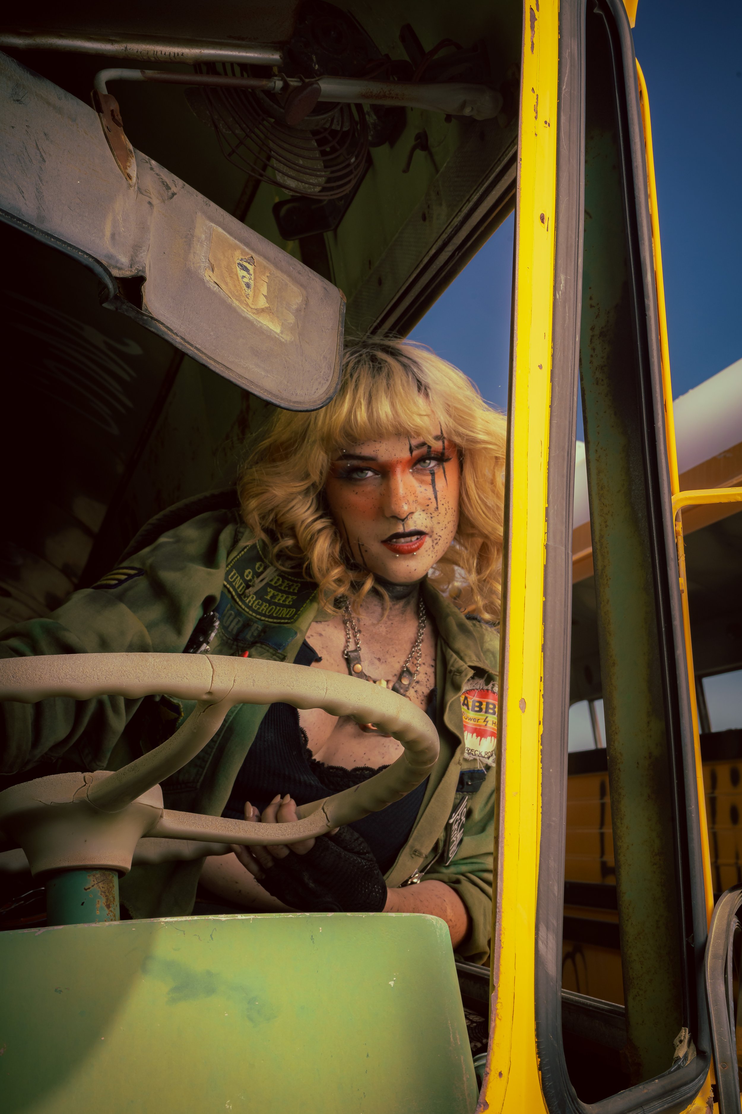 Person with makeup and blonde curly hair inside a green and yellow vehicle, possibly a school bus, looking out the window.