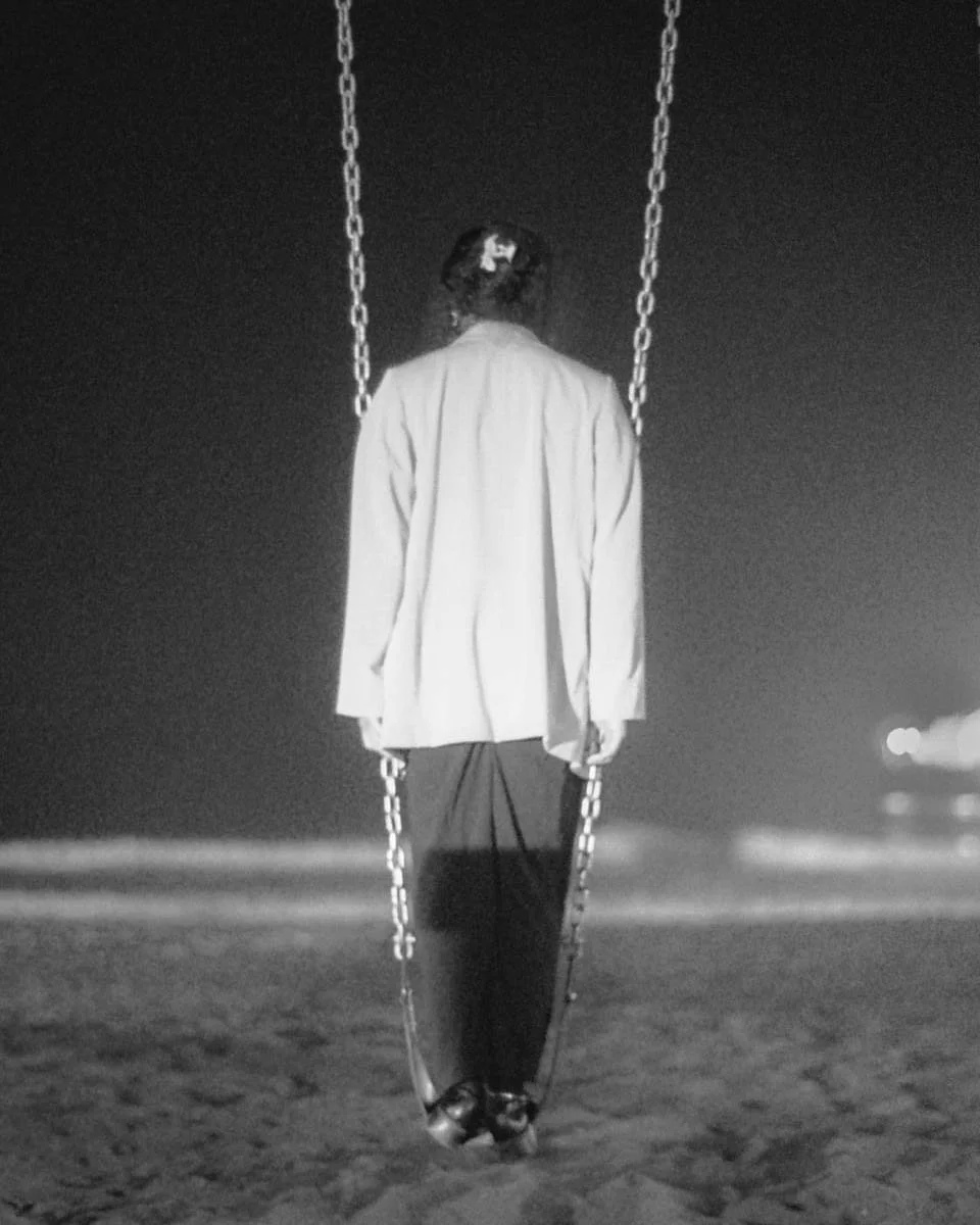 Person sitting on a swing at night on a beach, facing away from the camera. The photo is in black and white.