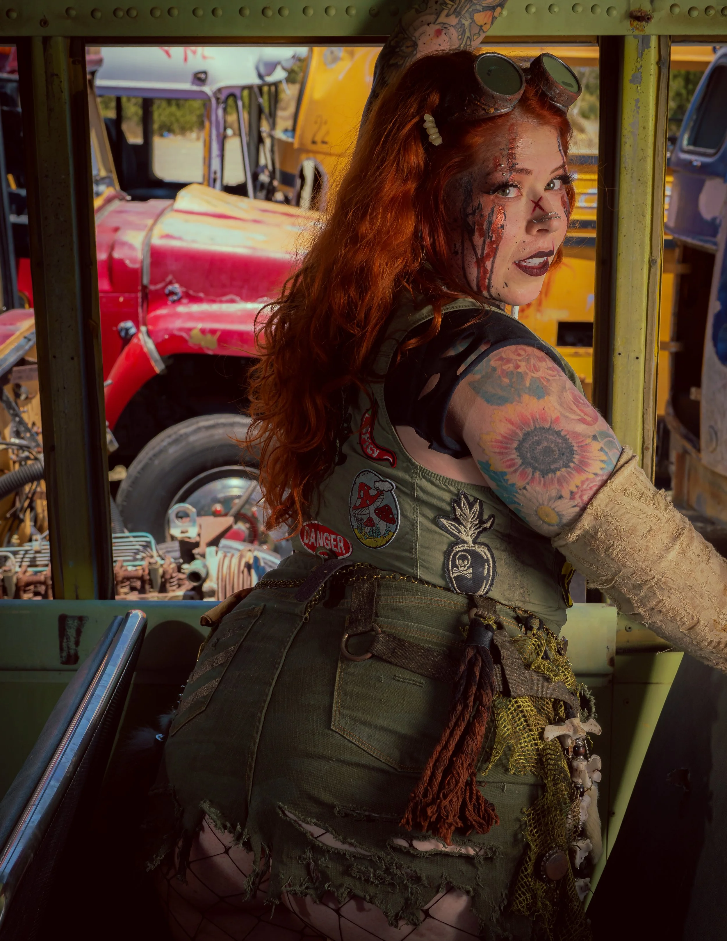 A woman with red hair and tattoos, wearing goggles on her head, leans out of a vehicle with a background of colorful, weathered vintage trucks.