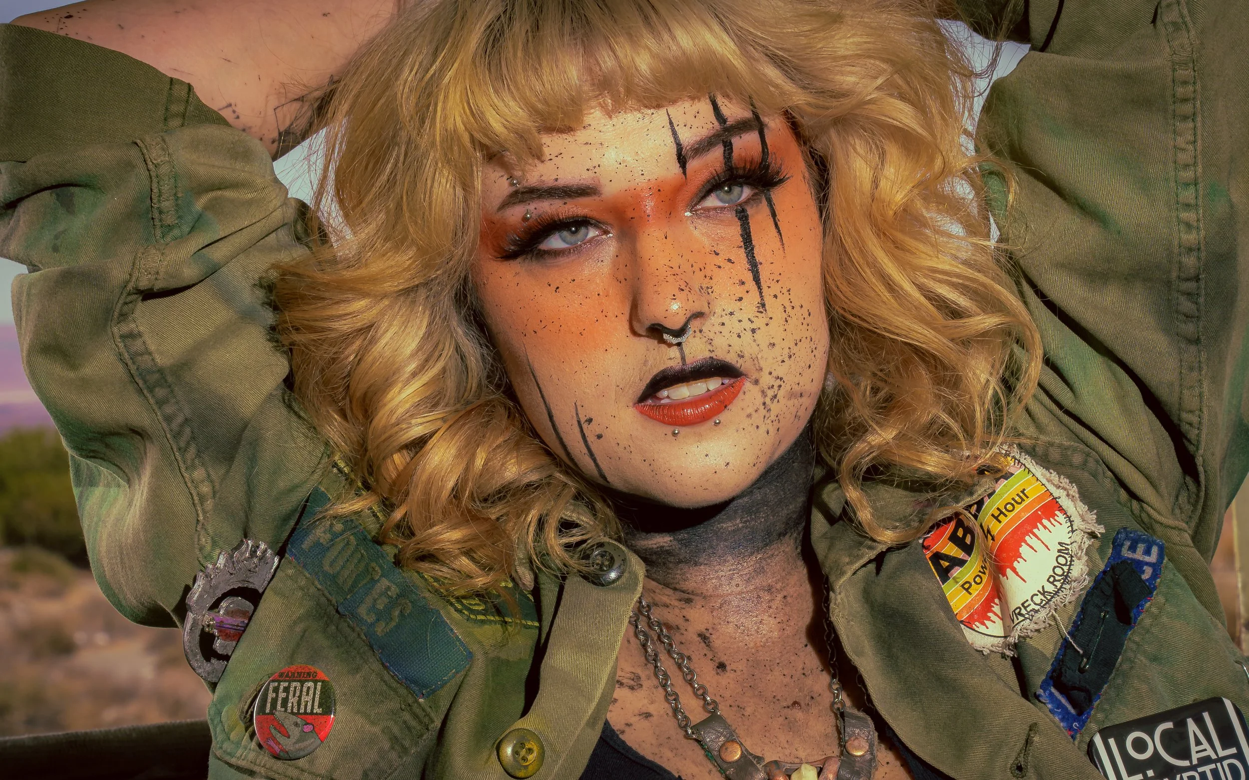 A woman with curly blonde hair, blue eyes, and distinctive makeup, wearing a green military jacket with patches and chains, outdoors with a blurred landscape in the background.