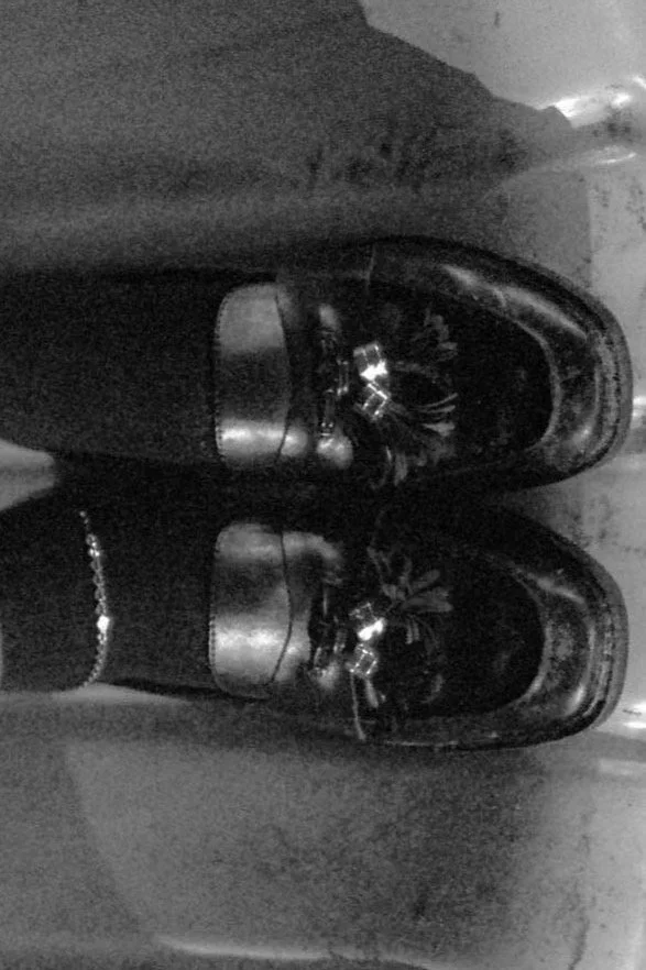 A pair of shiny black dress shoes worn with black pants and a black anklet, photographed from above.