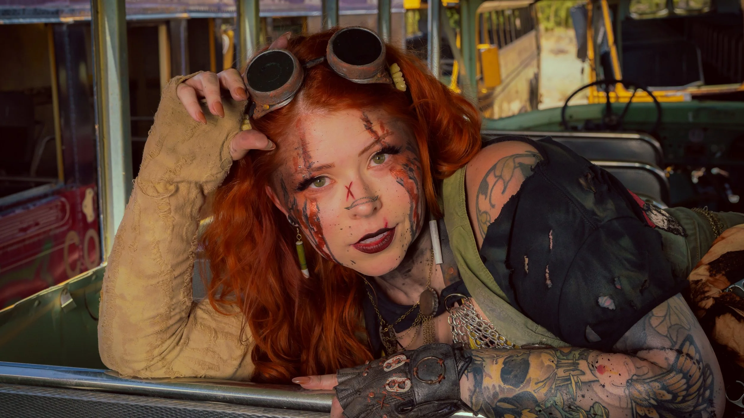 Woman with vibrant red hair, face tattoos, and dark makeup lying in an old vehicle, wearing goggles on her head and camouflage clothes with tattoos on her arms.