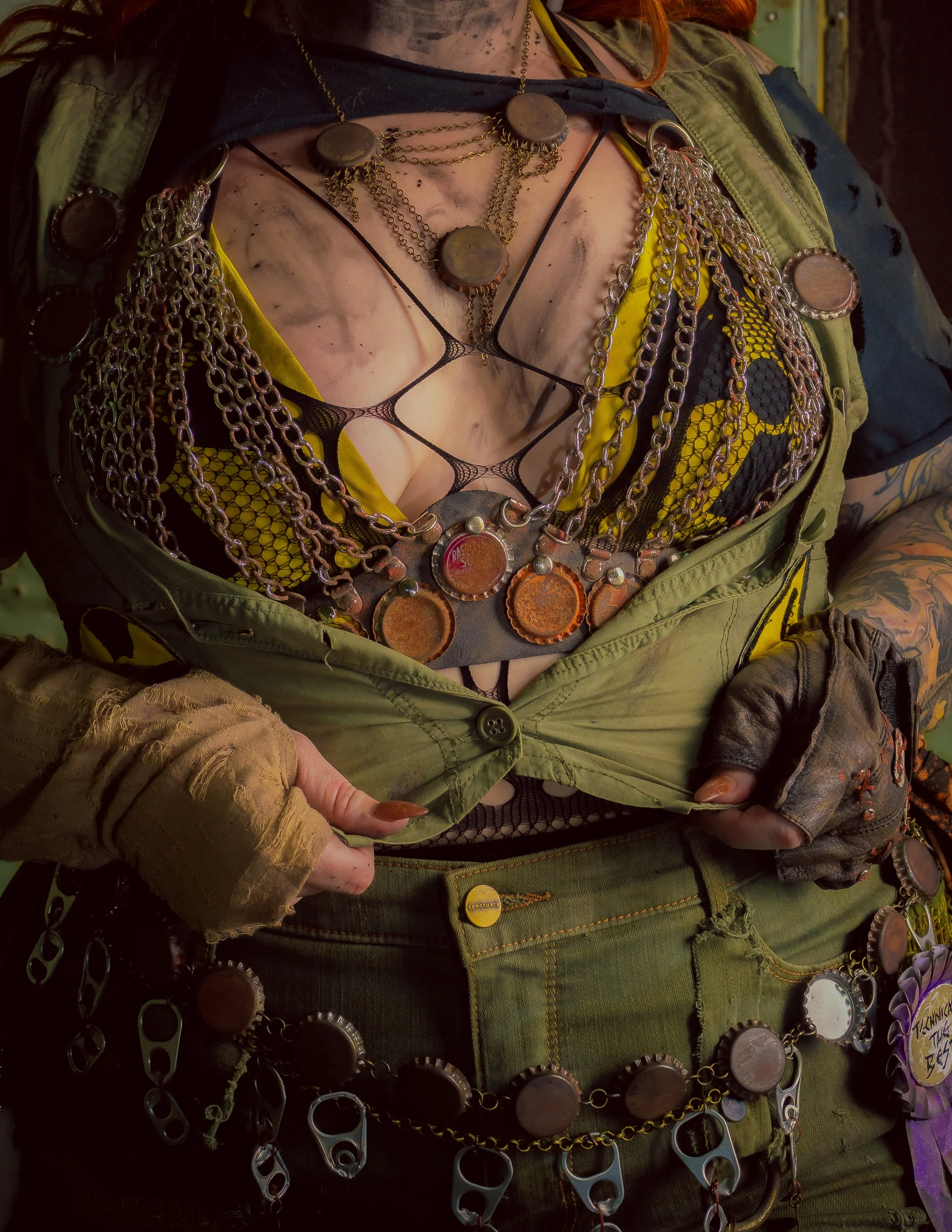 A person wearing a black mesh top, green jacket, and numerous layered necklaces, including chains and large metal medallions, with dirty makeup and tattoos. They are adjusting their jacket, with their hands in fingerless Gloves.