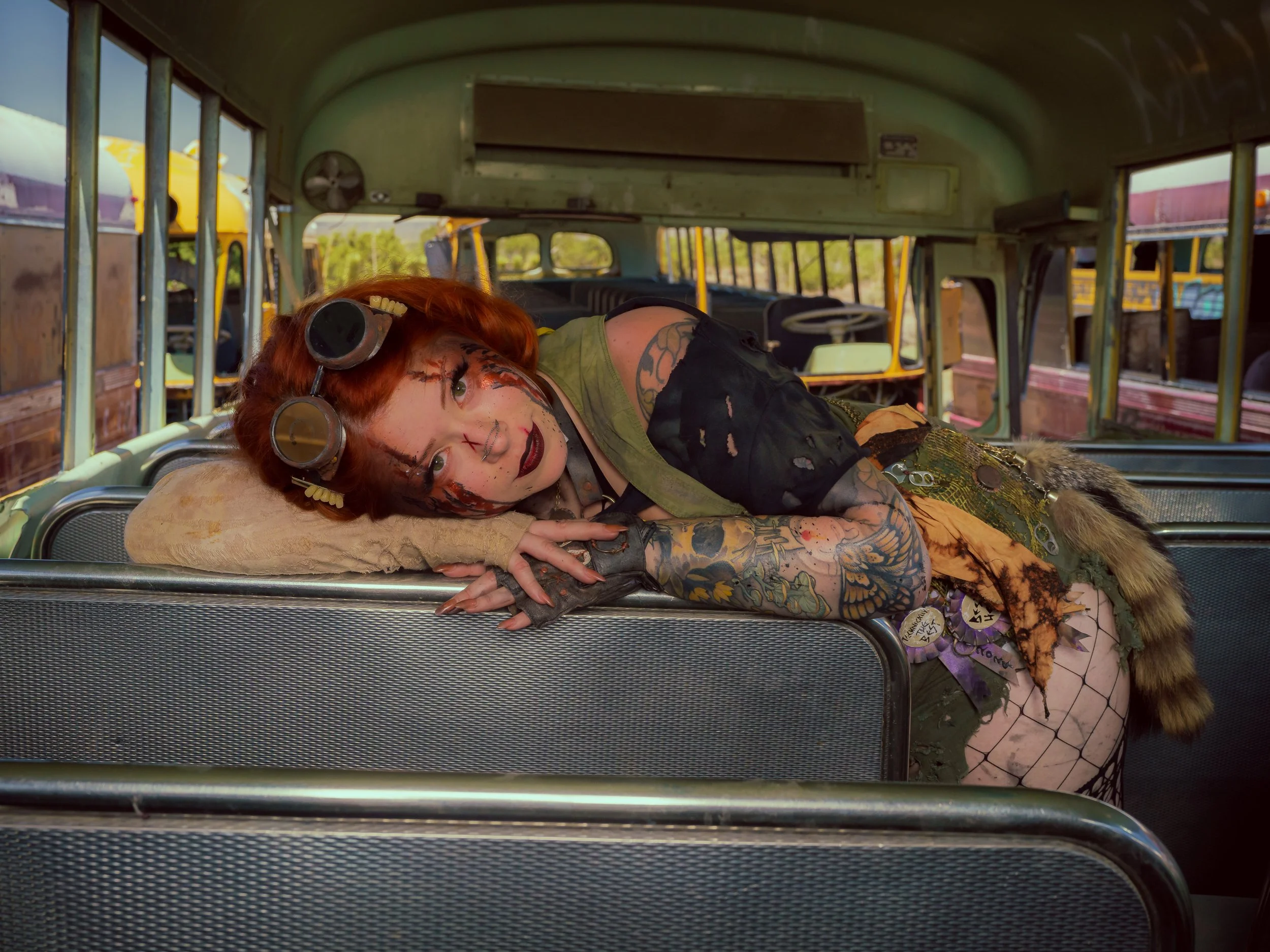 A woman with tattoos and makeup lying on her side on a vintage bus, wearing headphones and a costume with animal print and armor details.