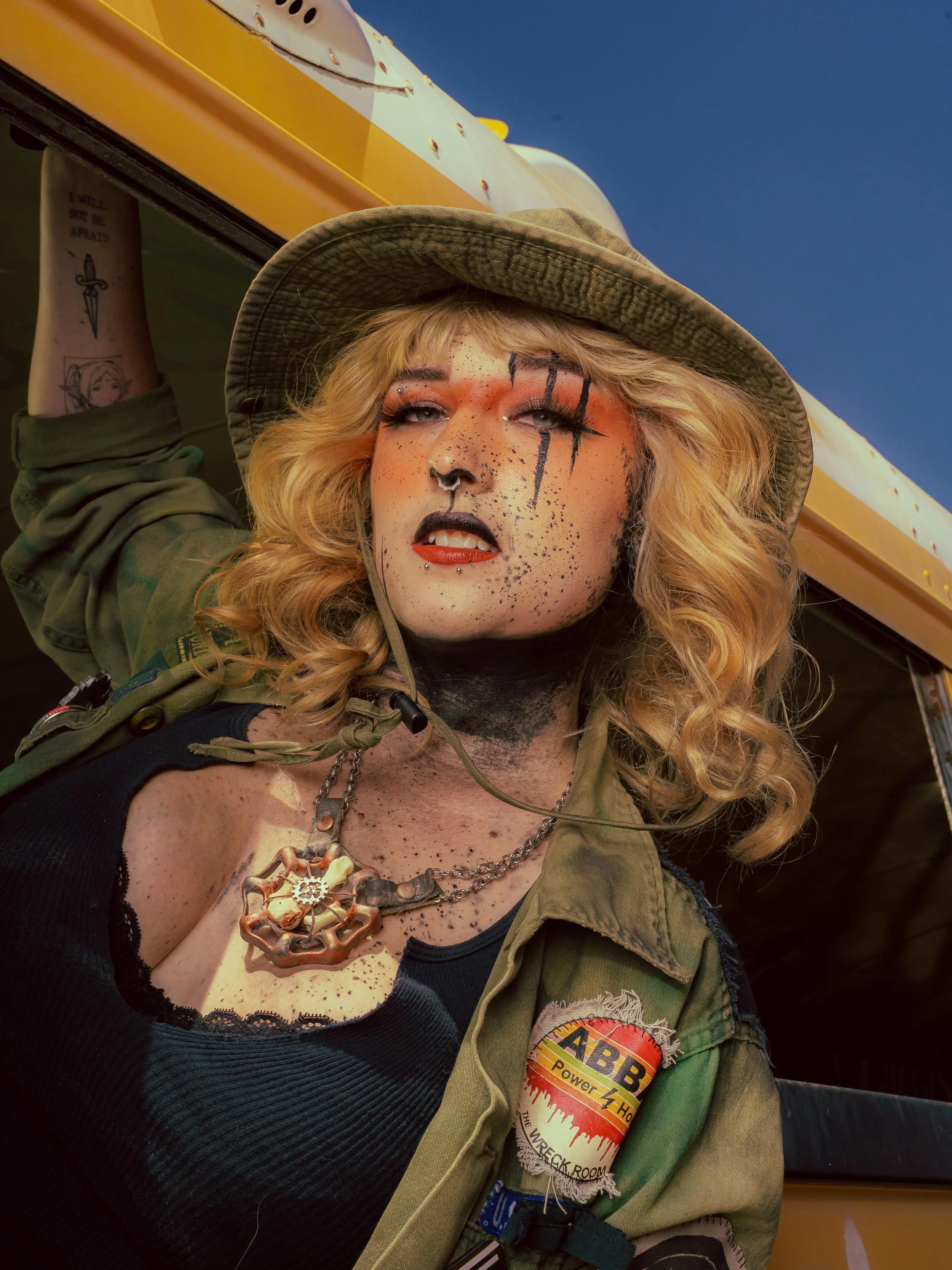 A person with elaborate makeup and tattoos, wearing a military-style jacket with patches and necklaces, posing in front of a yellow vehicle against a blue sky.