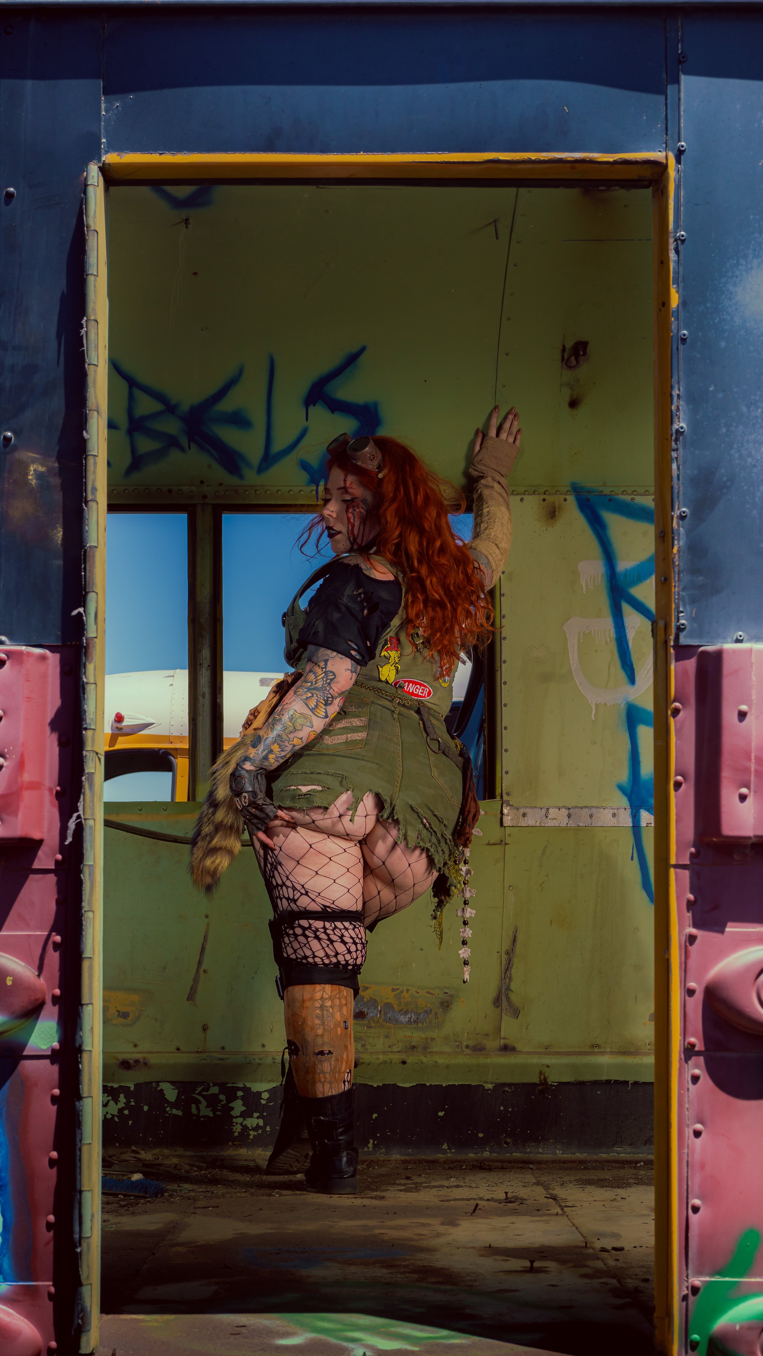 A woman with long curly red hair and tattoos on her arms and leg, dressed in distressed military-style clothing with fishnet stockings, stands in an old, graffiti-covered amusement park ride or container, looking back over her shoulder.