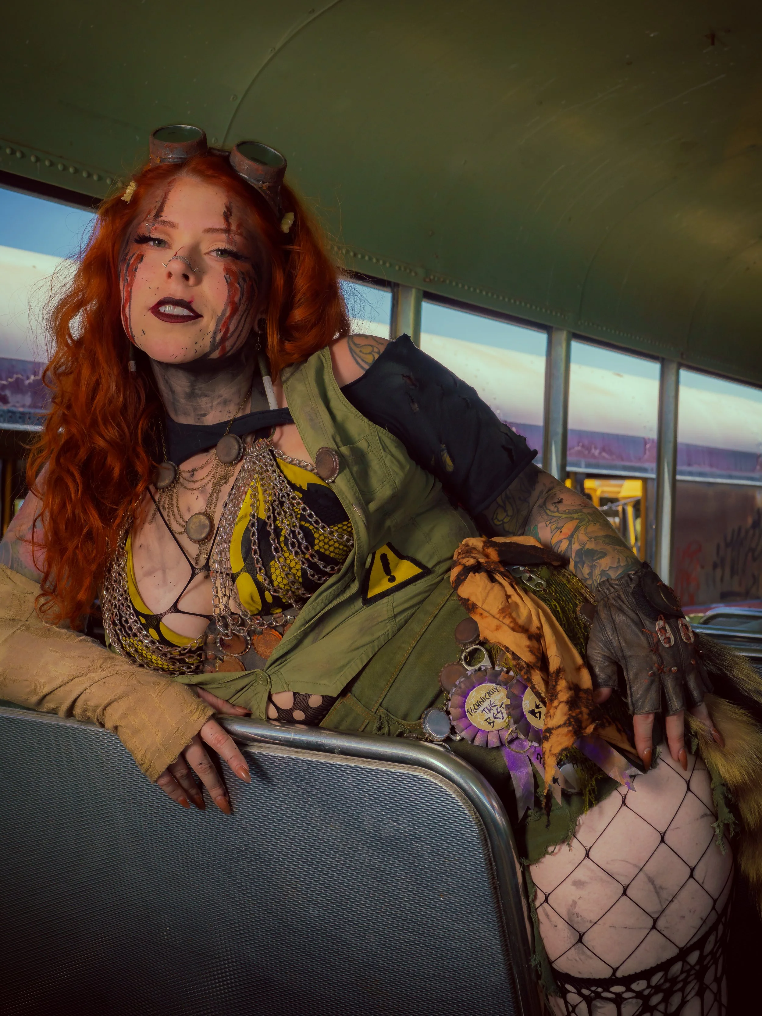 Woman with red curly hair, dressed in punk style, reclining on a bus seat, with makeup and tattoos, wearing a mix of clothing including military-style jacket, fishnet stockings, and layered jewelry.