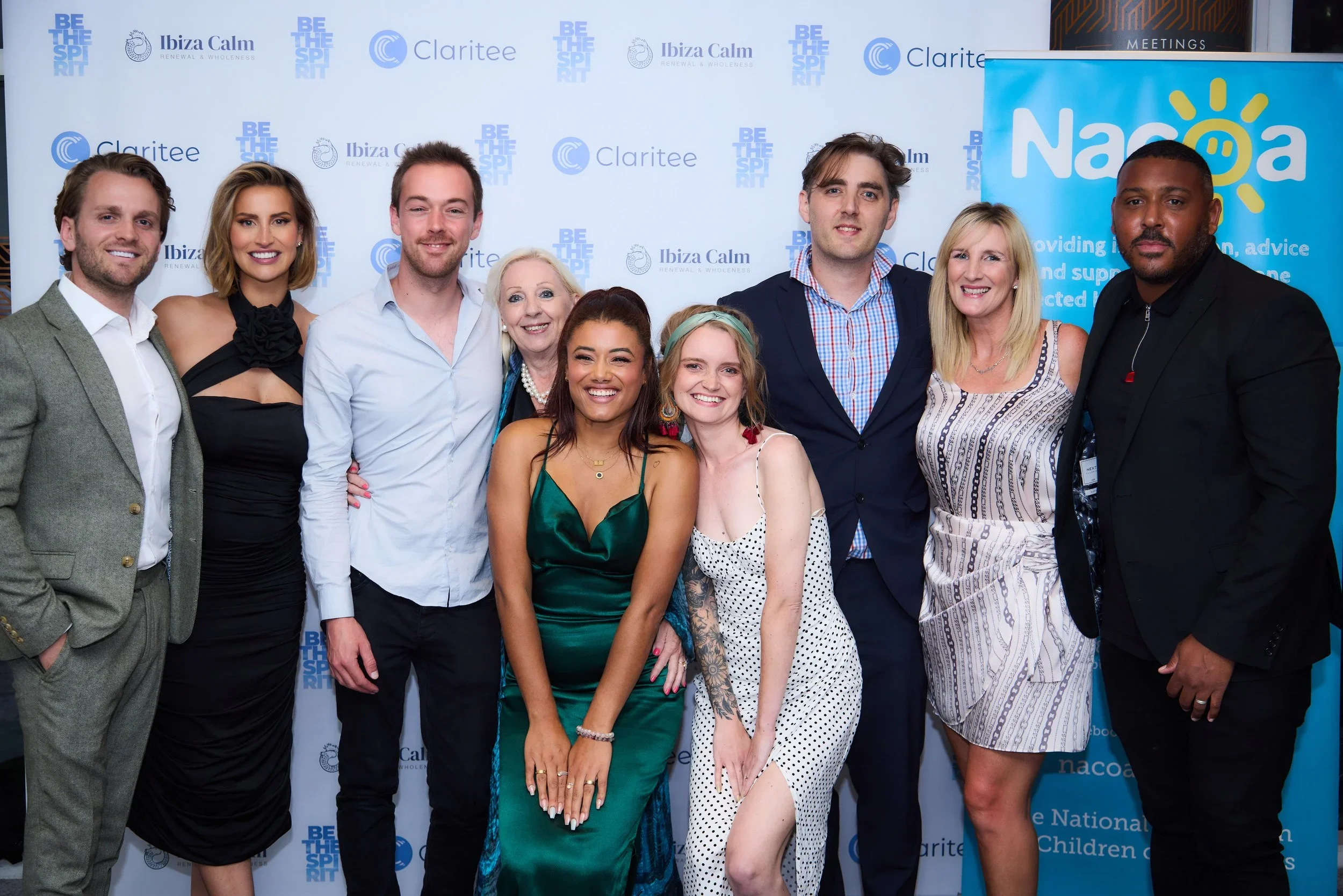 A group of ten people posing together at an event, standing in front of a backdrop with logos and a banner for Naco. They are dressed in formal and semi-formal attire, smiling for the photo.