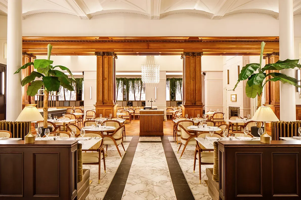 Elegant restaurant dining room with white marble floor, wooden columns, large leafy plants, and tables set for dining, with a chandelier hanging from the ceiling.