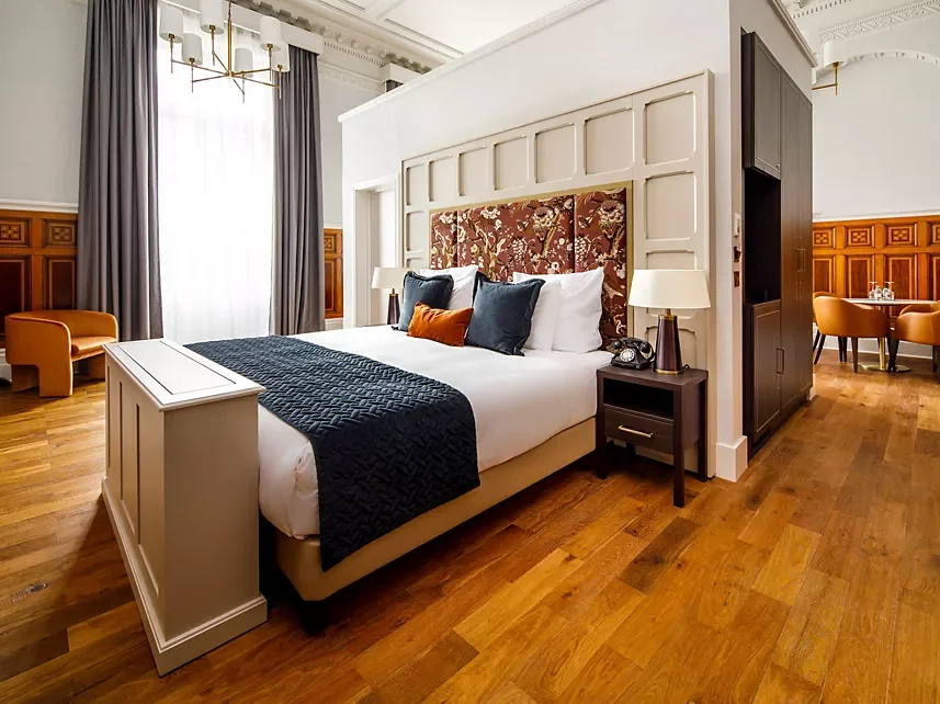 A hotel room with a large bed, pillows, and a padded headboard, in a room with wood floors, curtains, and a sitting area with a table and chairs.