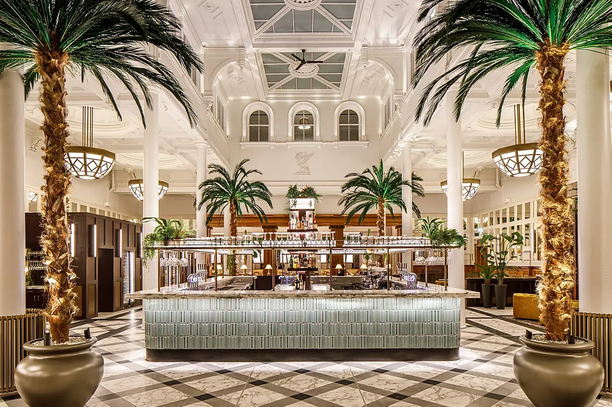 Elegant hotel lobby with a central bar, tall palm trees, white columns, high arched windows, and modern lighting fixtures.