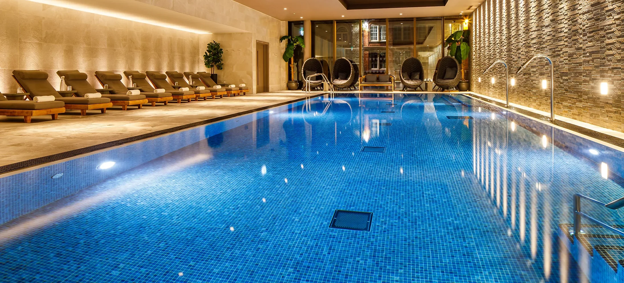 Indoor swimming pool with blue tiles, lounge chairs on the left, wicker chairs and large potted plants at the far end, and stone accent wall on the right. Warm lighting creates a relaxing atmosphere.