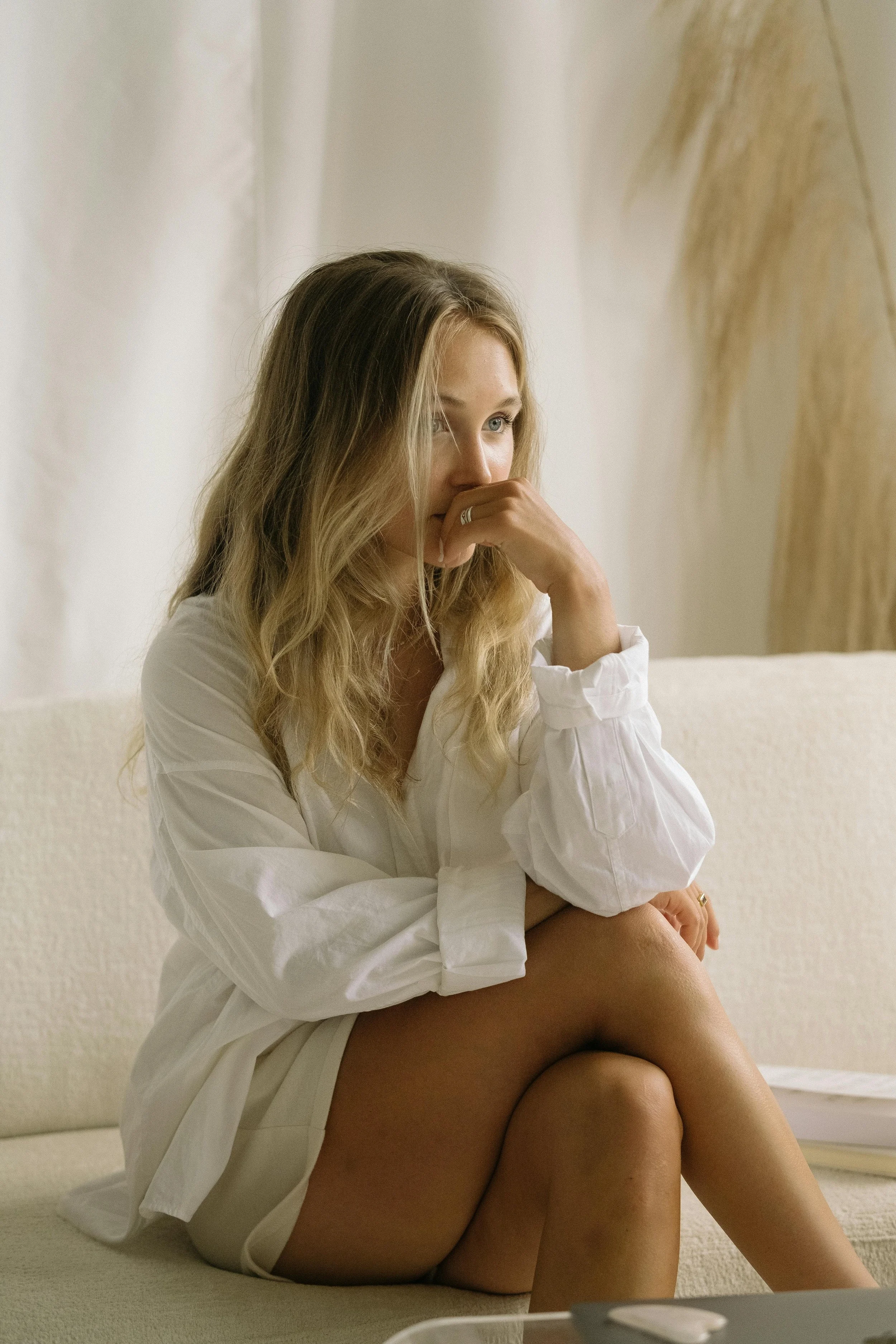 A woman with long blonde hair sitting on a beige sofa, wearing a white shirt, with her right hand resting on her knee and her left hand near her face, looking contemplative.
