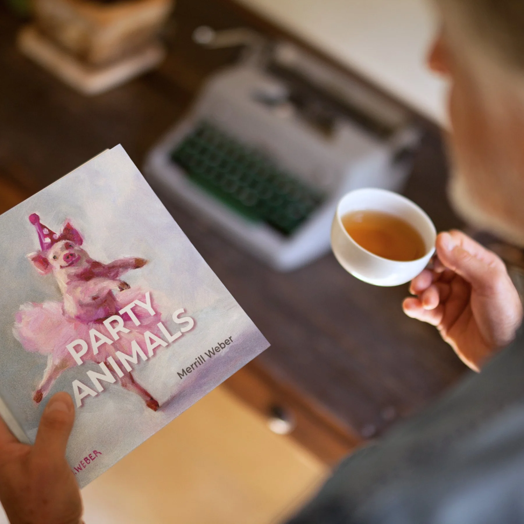 Person holding Party Animals art book beside a cup of tea and vintage typewriter in warm natural light.
